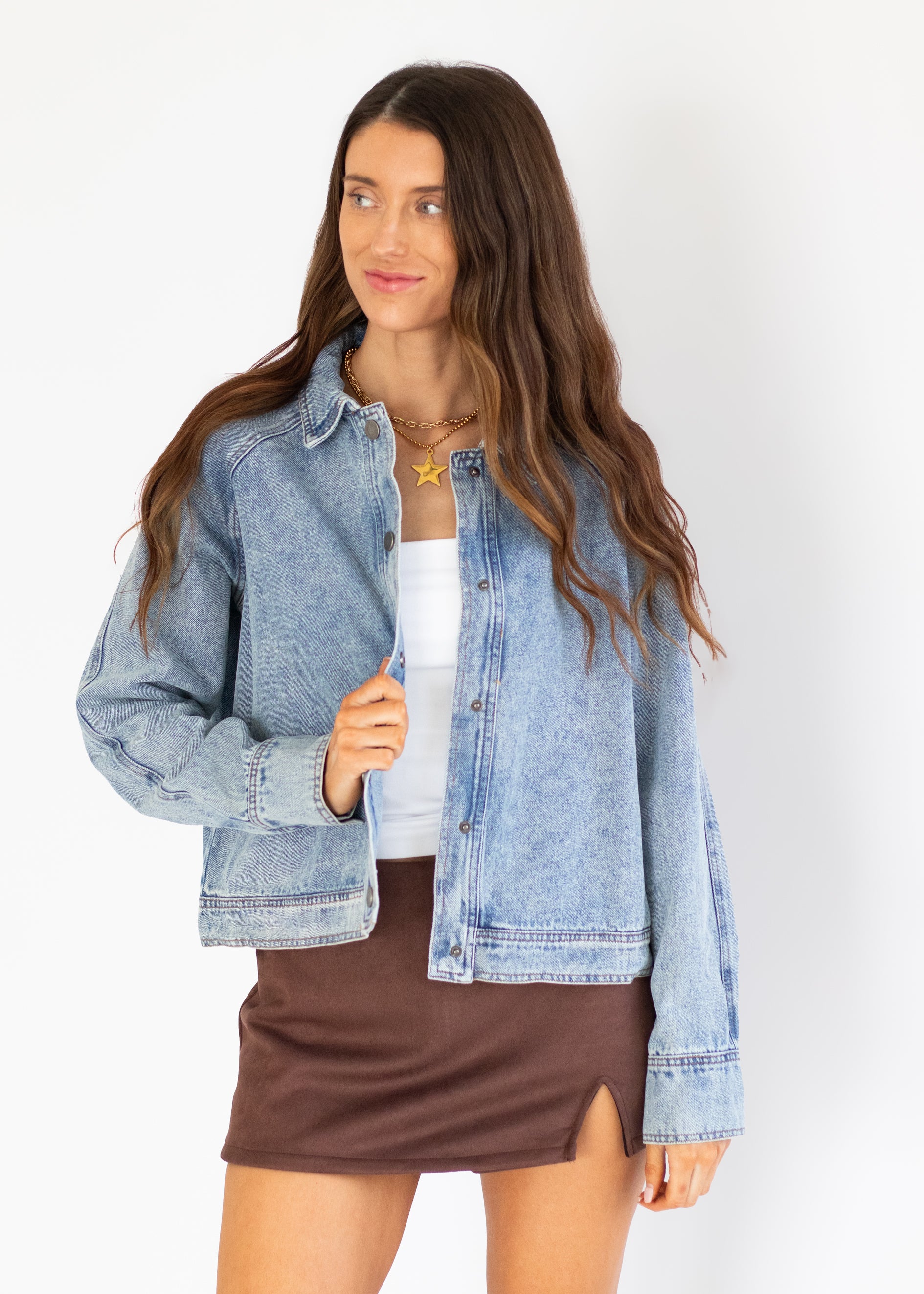 A woman modeling a light-wash blue denim jacket over a white top and brown mini skirt, standing against a plain white studio background.