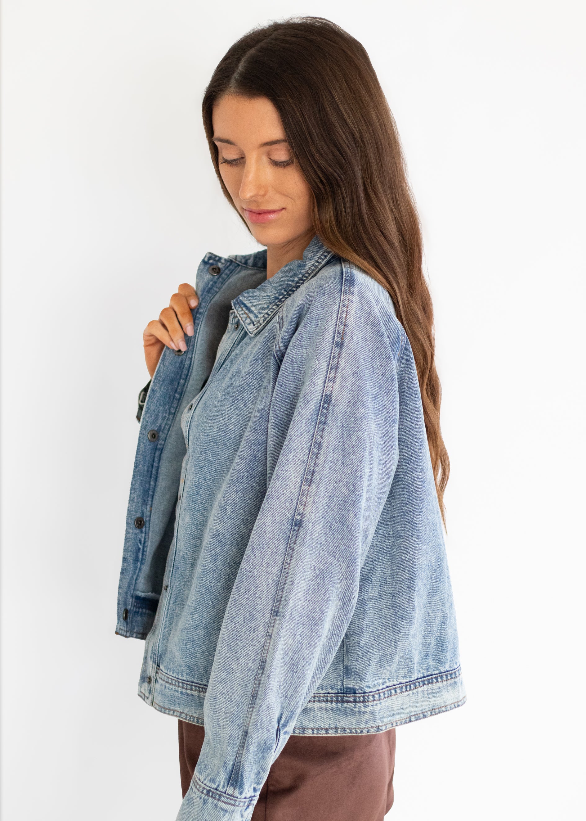 A woman wearing an oversized light-wash blue denim jacket over a gray top and brown pants, photographed against a plain white studio background.