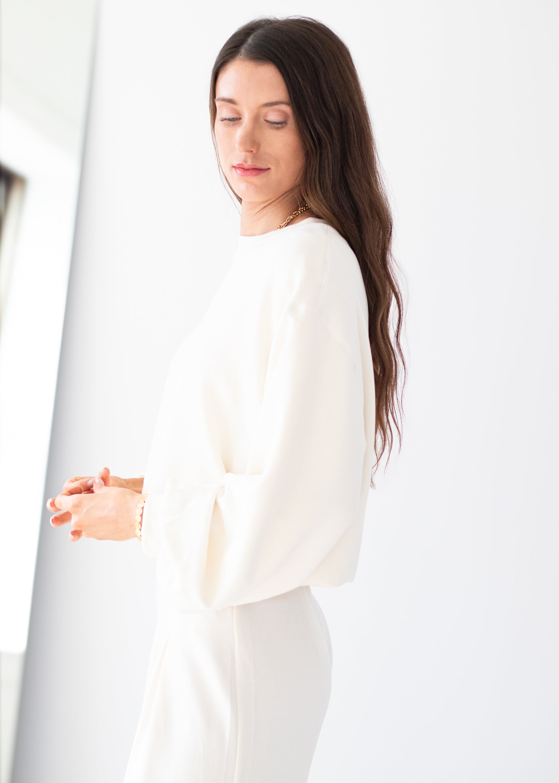 A woman with long dark brown hair is shown in profile wearing a white long-sleeve top and matching white bottoms, standing against a bright white background.