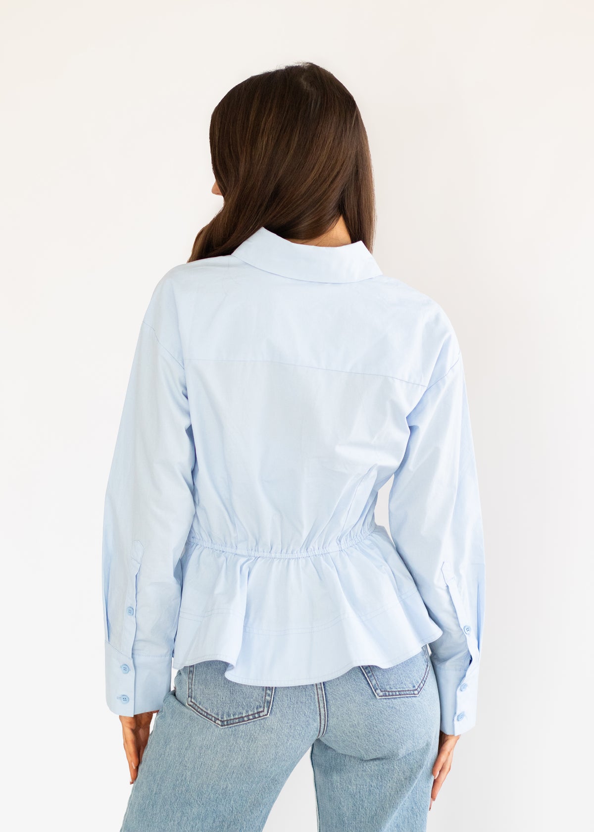 A woman viewed from behind wearing a light blue long-sleeve button-up shirt with a gathered peplum waist and light-wash blue jeans, set against a plain off-white background.