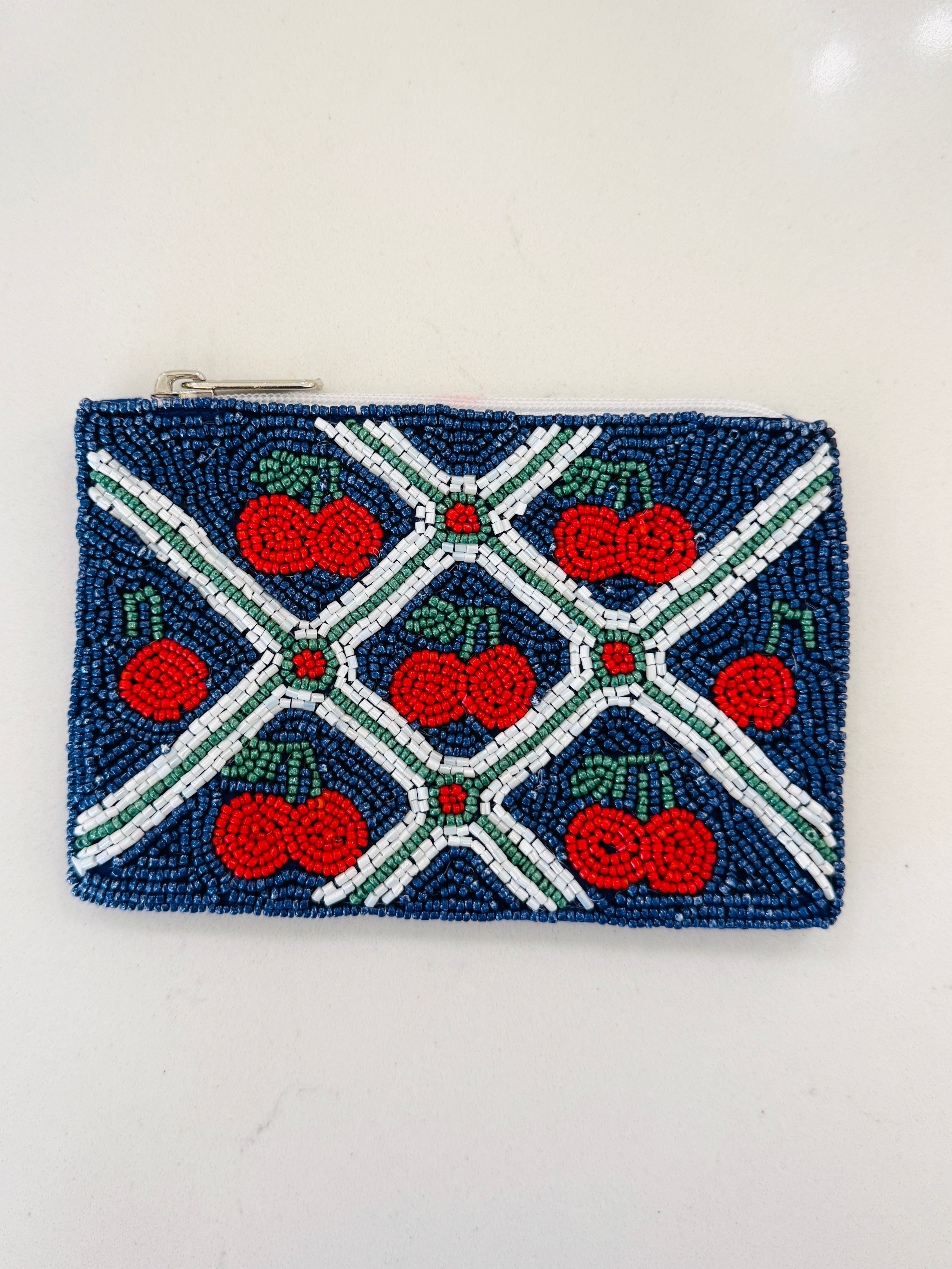 A small rectangular beaded coin purse with a zipper closure, featuring a navy blue background, red cherries with green stems, and white and green beaded geometric lines.