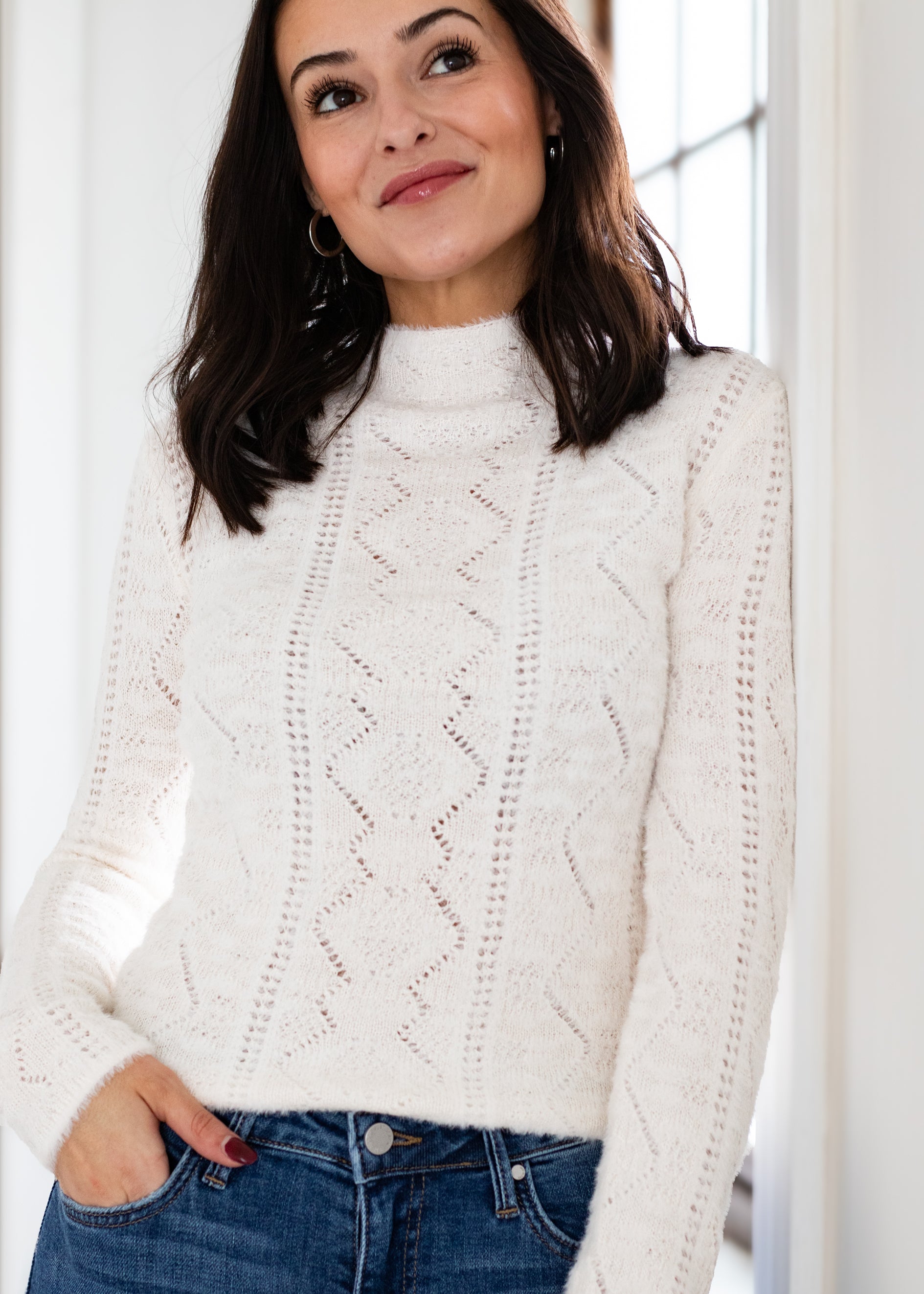 Woman wearing a white cable knit sweater with a blurred indoor background