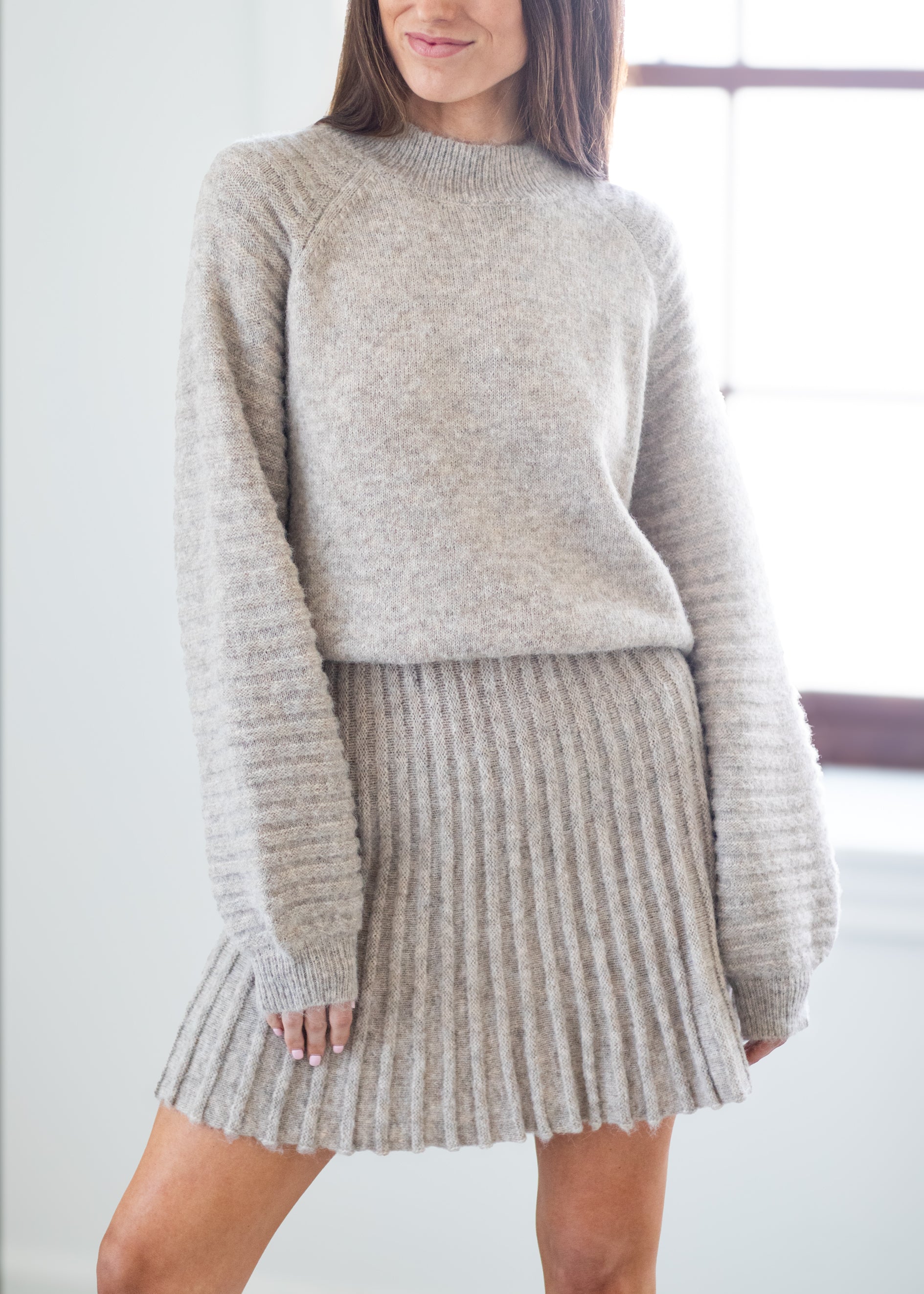 A woman wearing a light beige knit sweater and matching ribbed knit pleated skirt, standing in a bright indoor setting.
