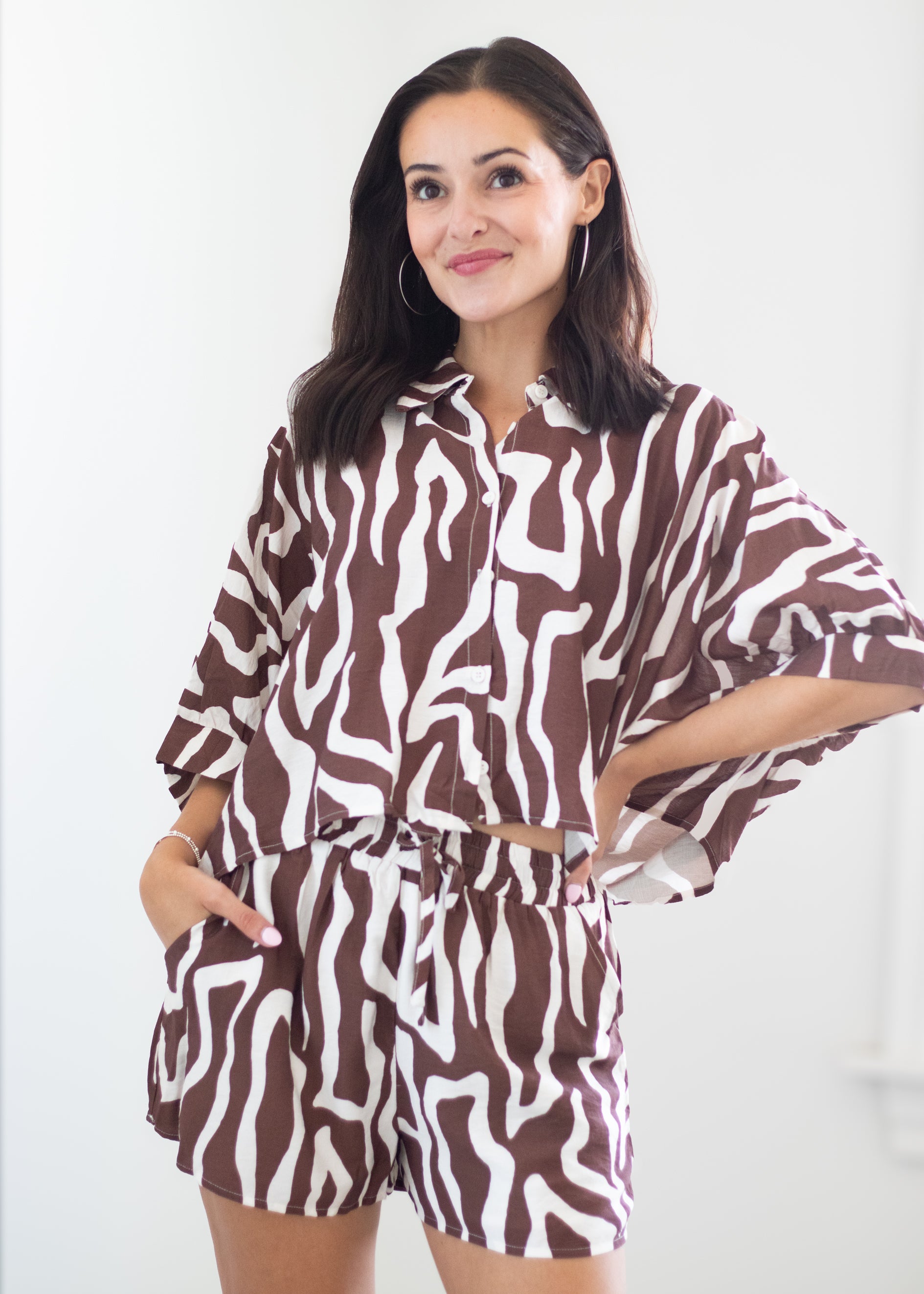 A woman modeling a matching brown and white zebra-print two-piece set consisting of a cropped button-up shirt and shorts, set against a plain white background.