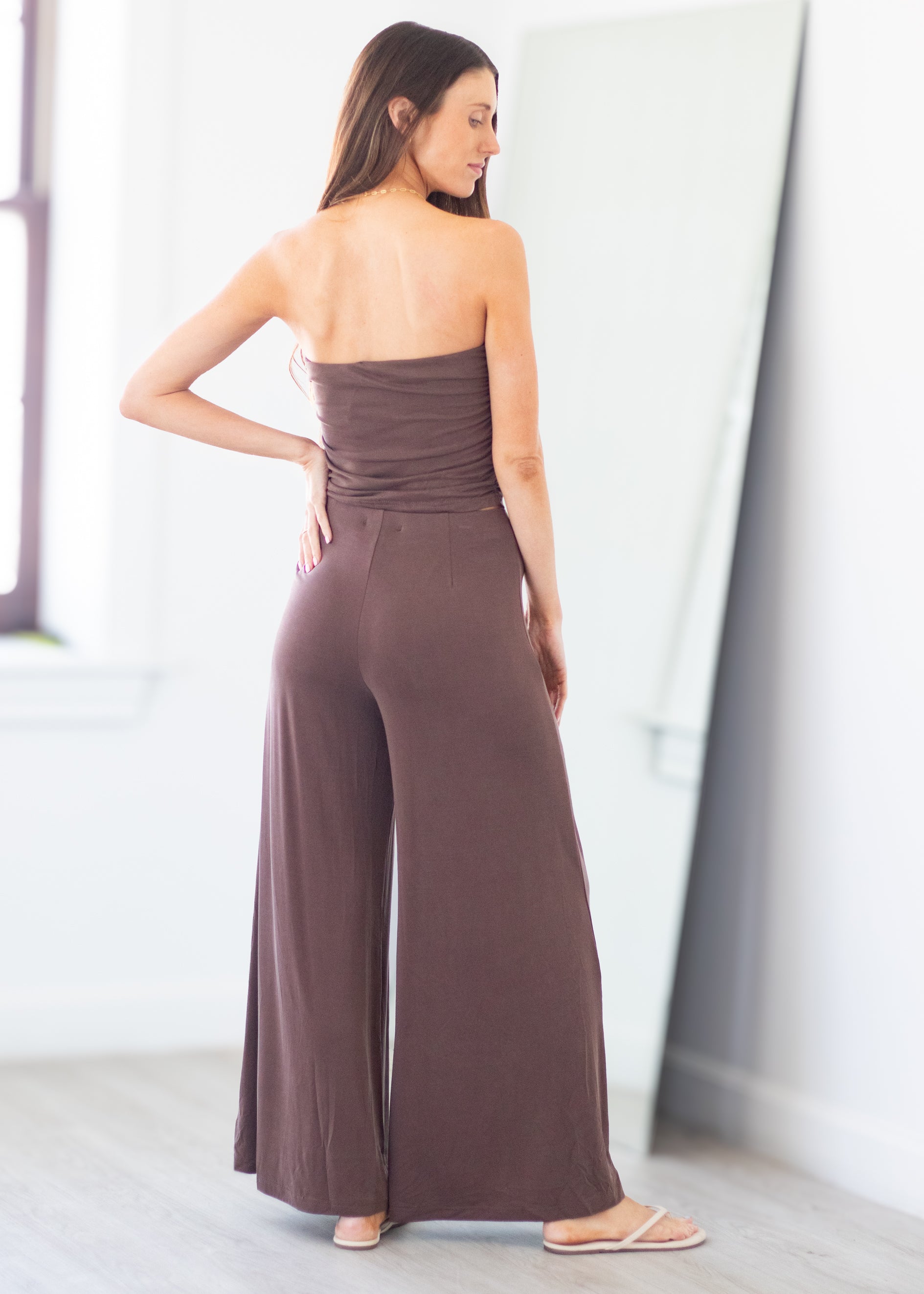 A woman wearing a strapless wide-leg jumpsuit in a muted brownish-purple shade, standing in a bright indoor room with a mirror in the background.