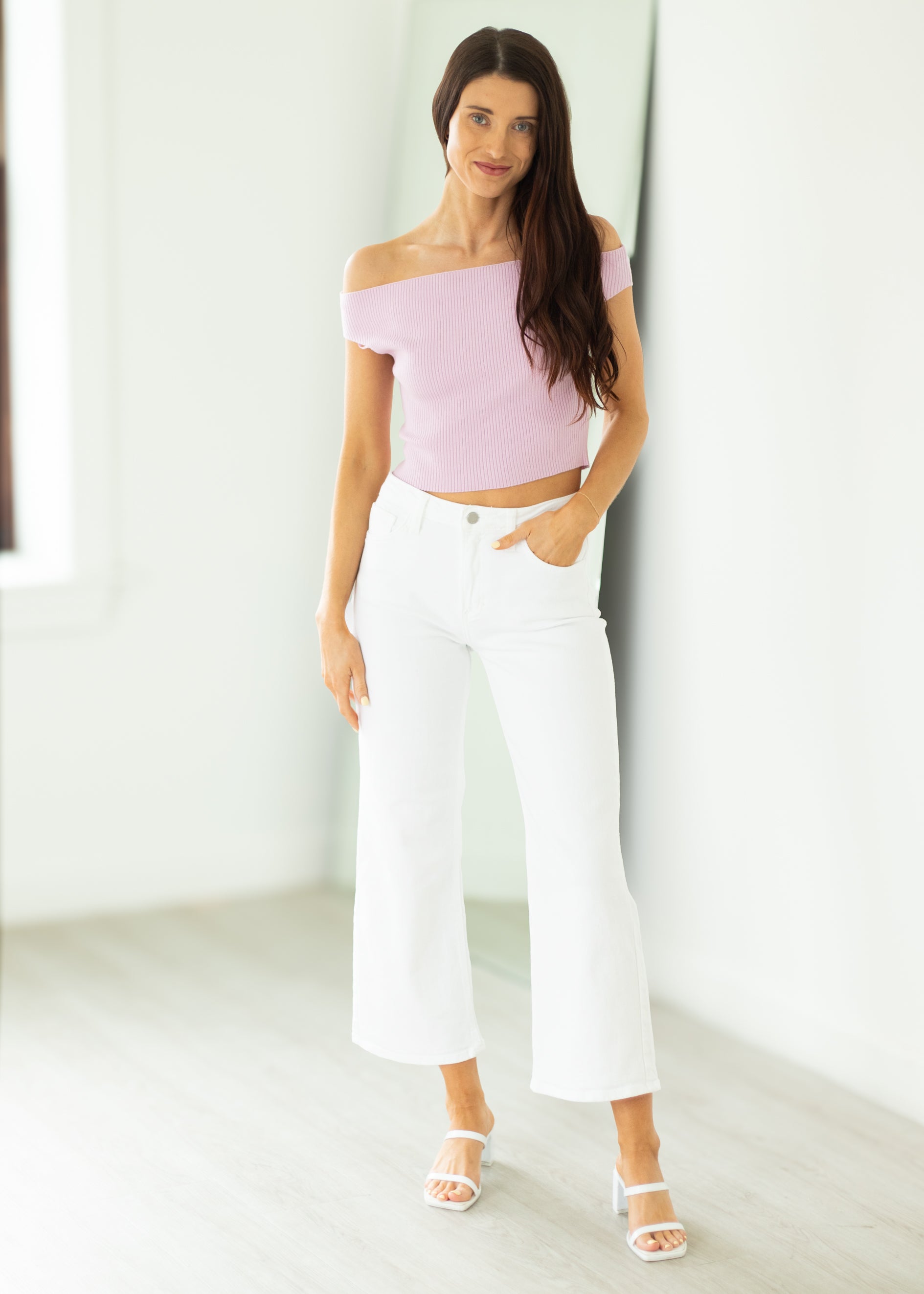 Woman wearing a light purple off-shoulder top and white pants in a minimalistic setting.