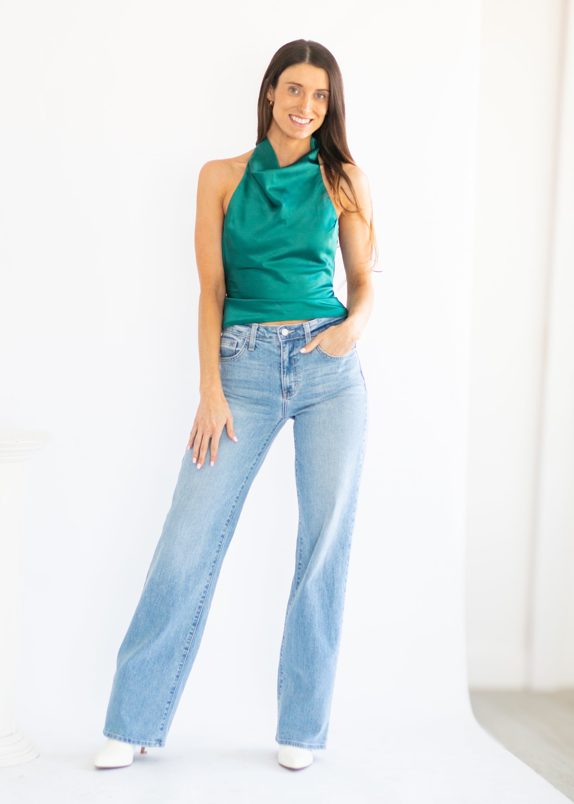 A woman modeling a sleeveless emerald green satin halter top and light-wash wide-leg blue jeans against a bright white studio background.