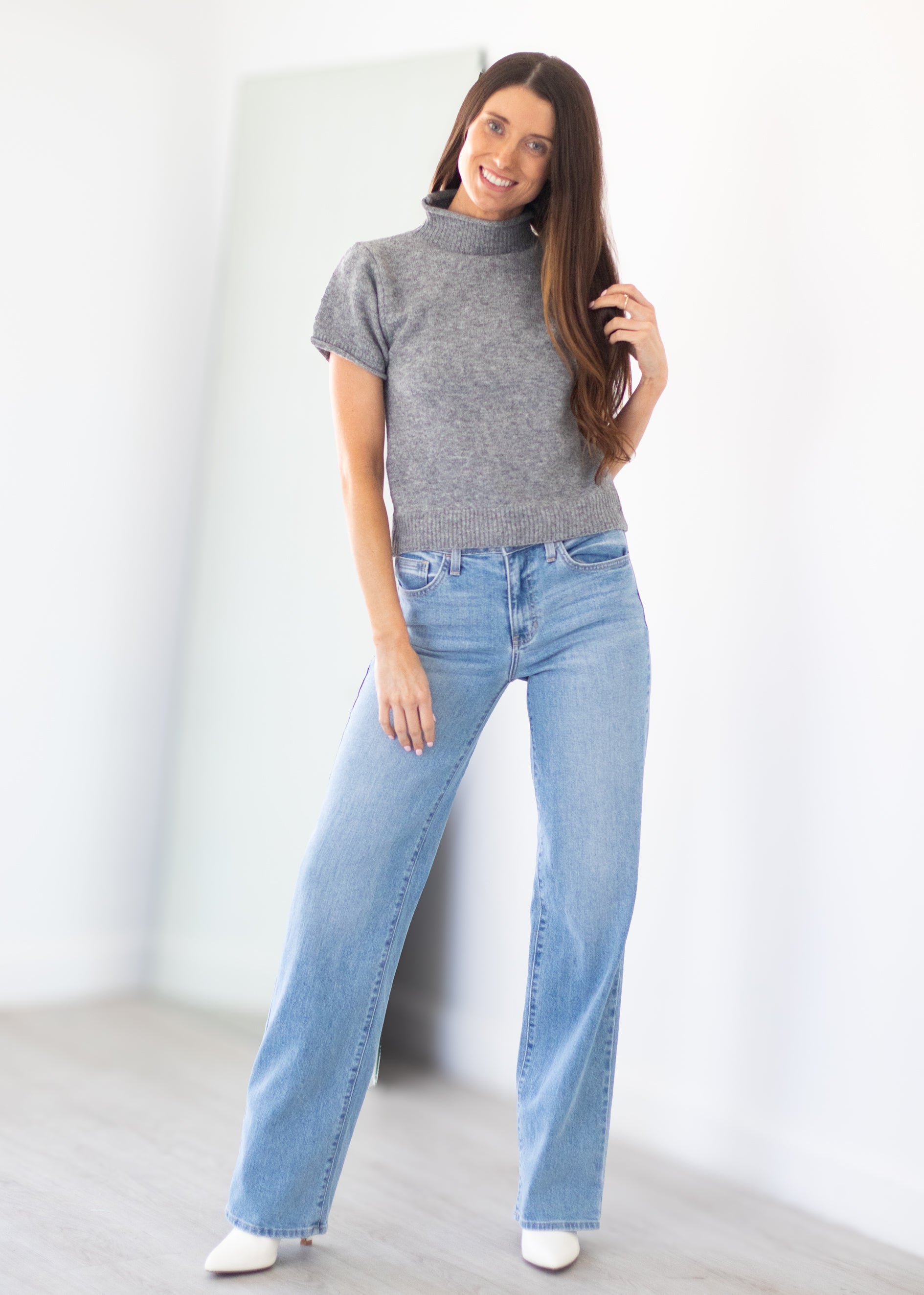 A woman modeling a gray short-sleeve mock neck knit top and light-wash blue wide-leg jeans, standing in a bright, minimalist room with white walls and light wood flooring.