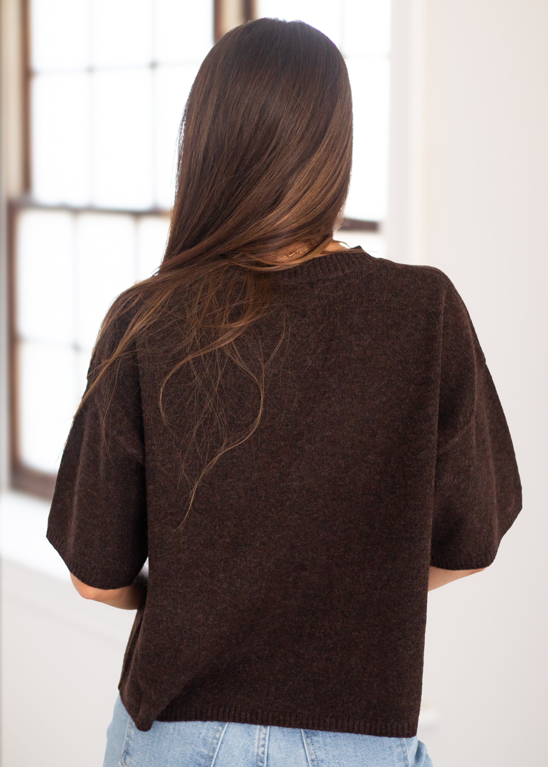 A woman viewed from behind wearing a dark brown short-sleeve knit sweater and light blue jeans, standing in a bright indoor setting near a window.