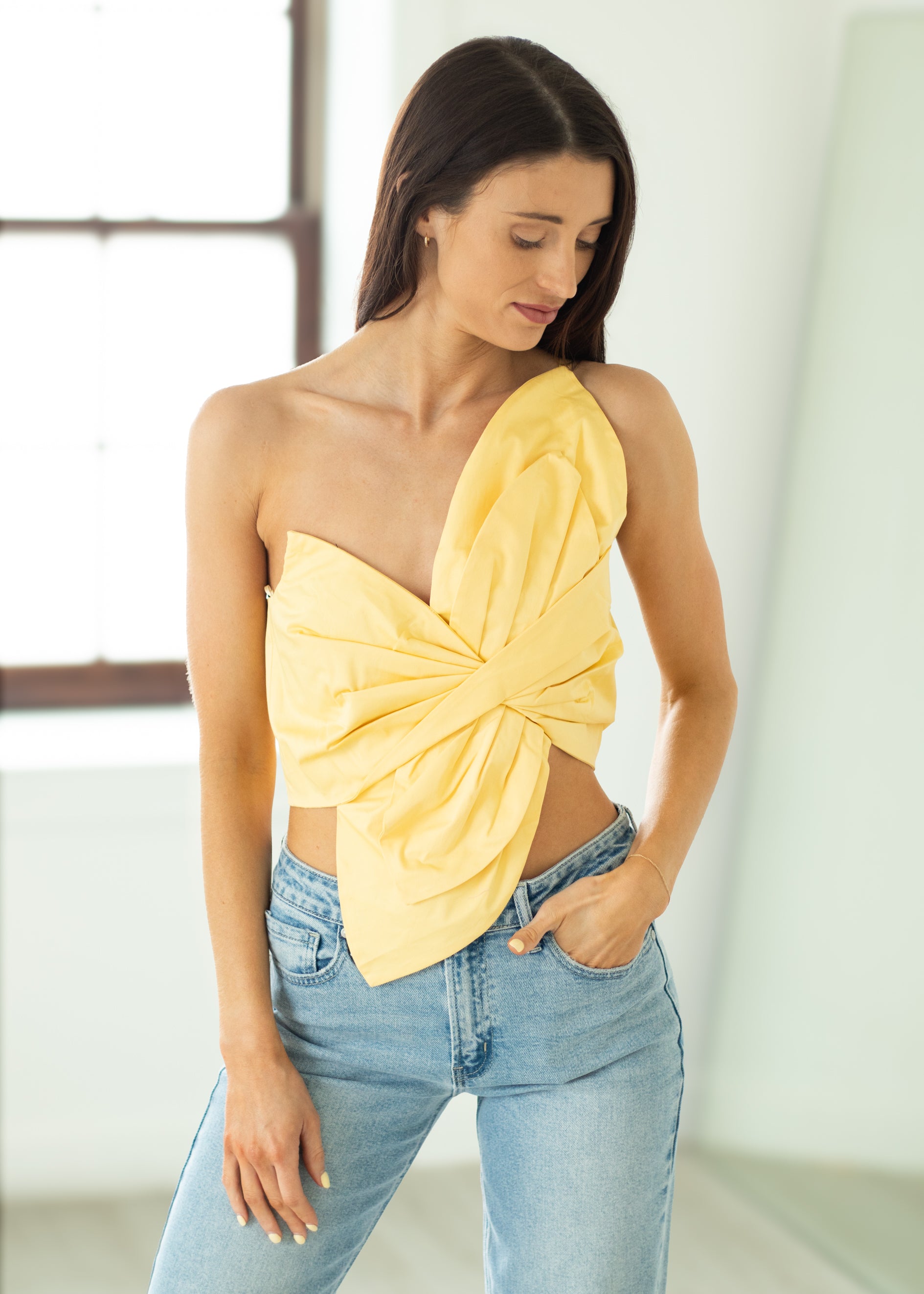 Woman wearing a yellow one-shoulder top and blue jeans indoors.