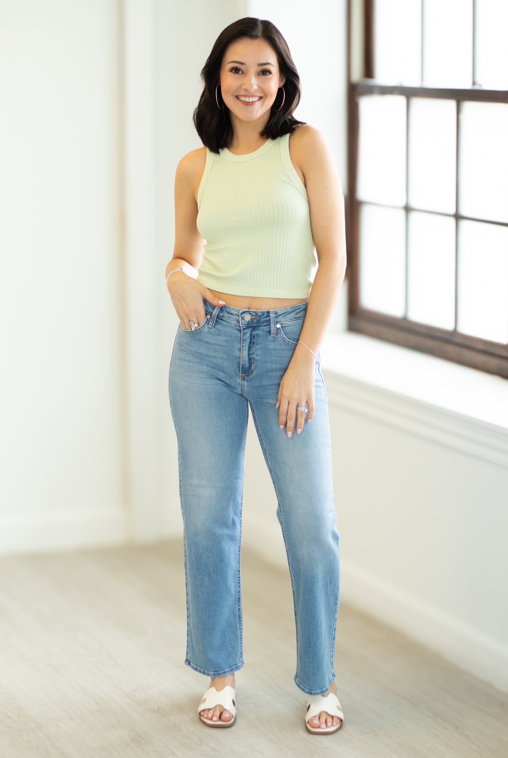 Woman wearing a light green sleeveless top and blue jeans standing in a room with a large window.