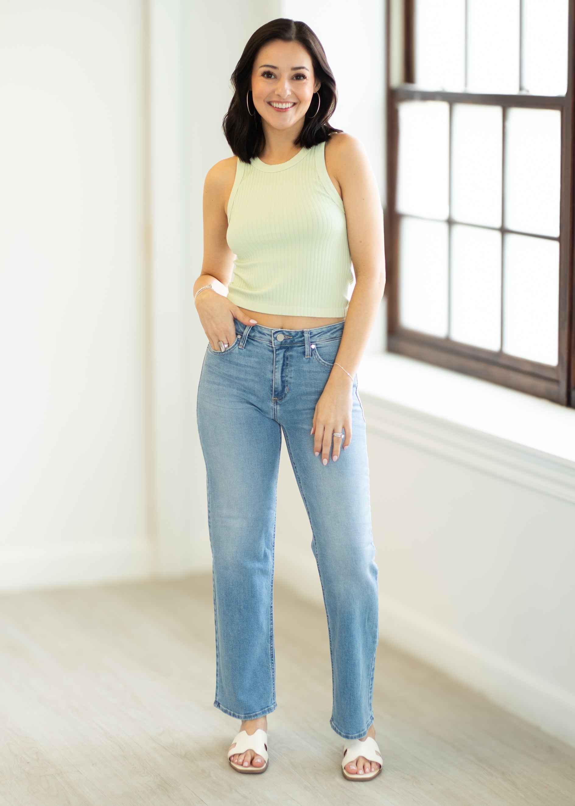 Woman wearing a light green sleeveless top and blue jeans standing in a room with a large window.