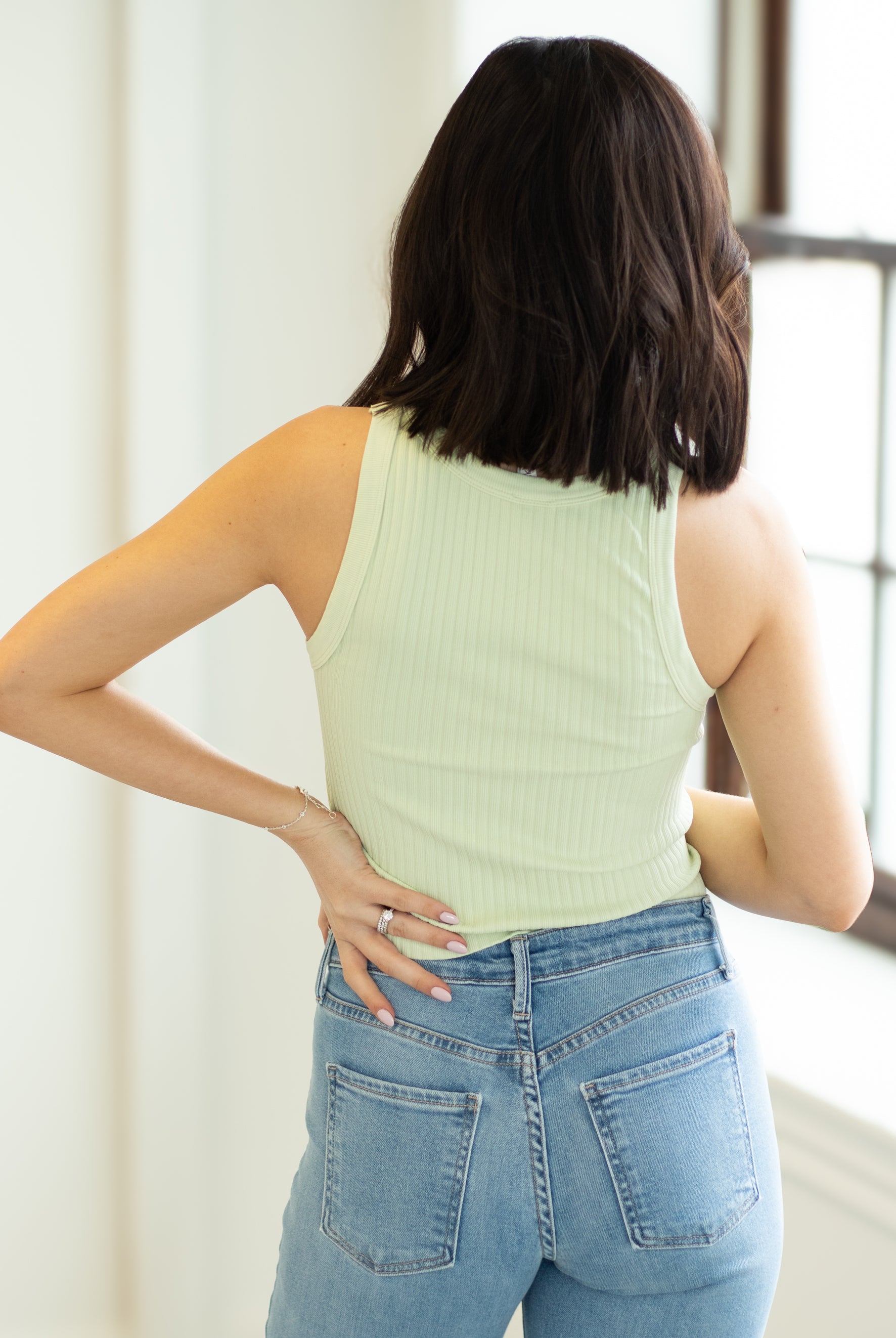 Woman wearing a light green sleeveless top and blue jeans, standing indoors.