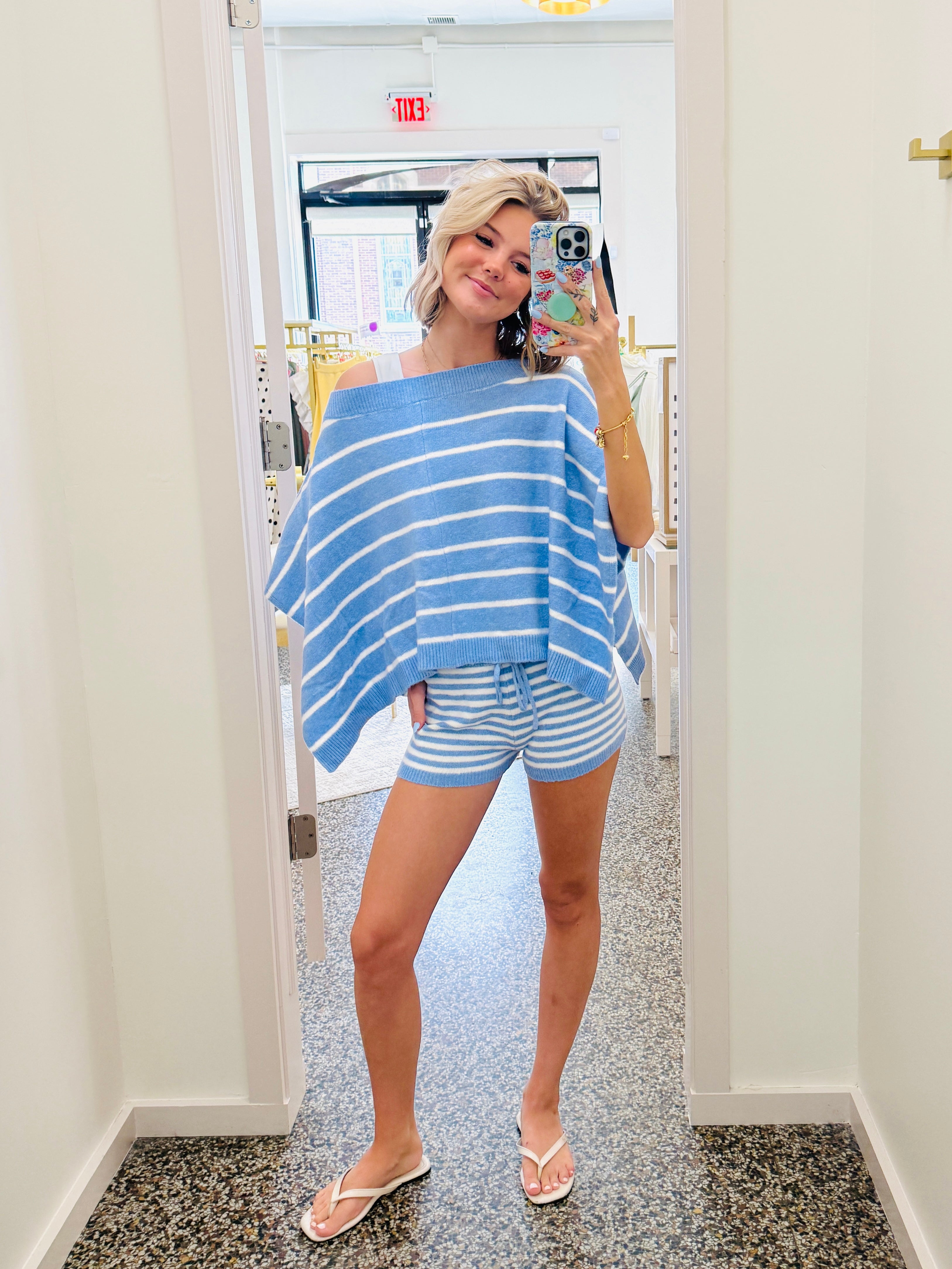 A woman takes a mirror selfie in a boutique wearing a light blue and white striped off-the-shoulder poncho top and matching striped shorts.