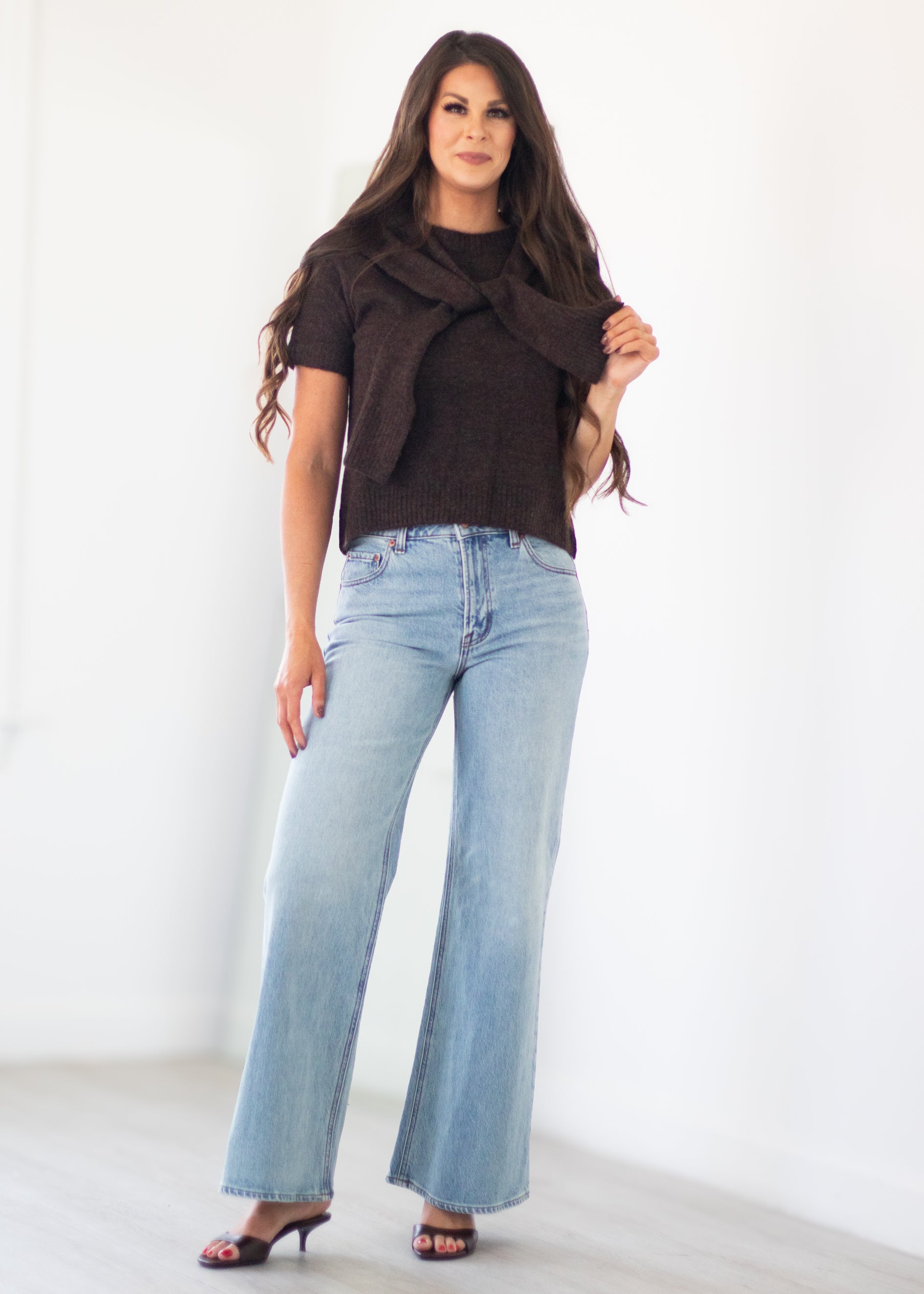 A woman modeling a dark brown knit wrap top and light-wash blue wide-leg jeans against a bright white studio background.