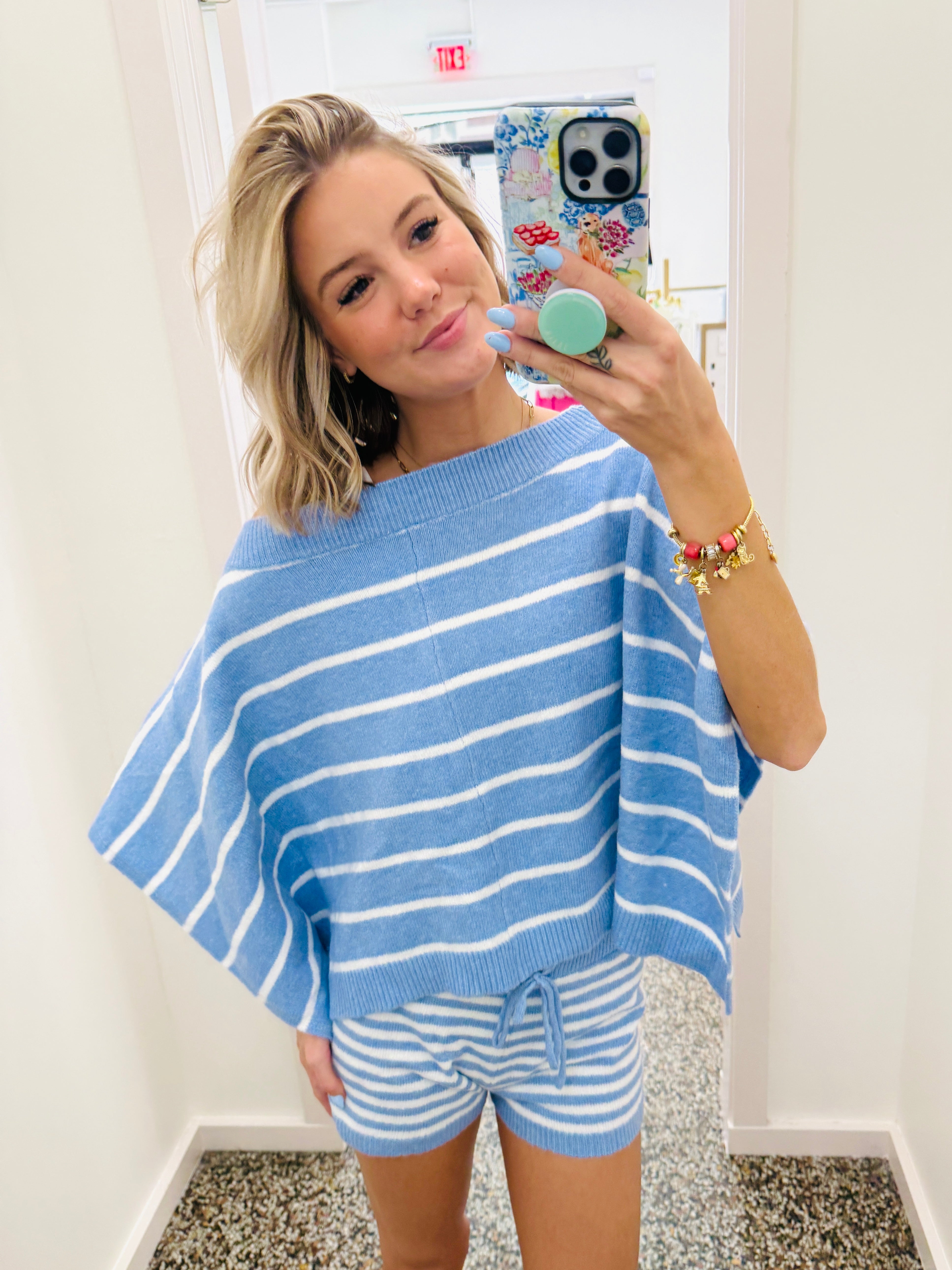 A woman takes a mirror selfie wearing a light blue and white horizontally striped knit poncho sweater and matching striped shorts.