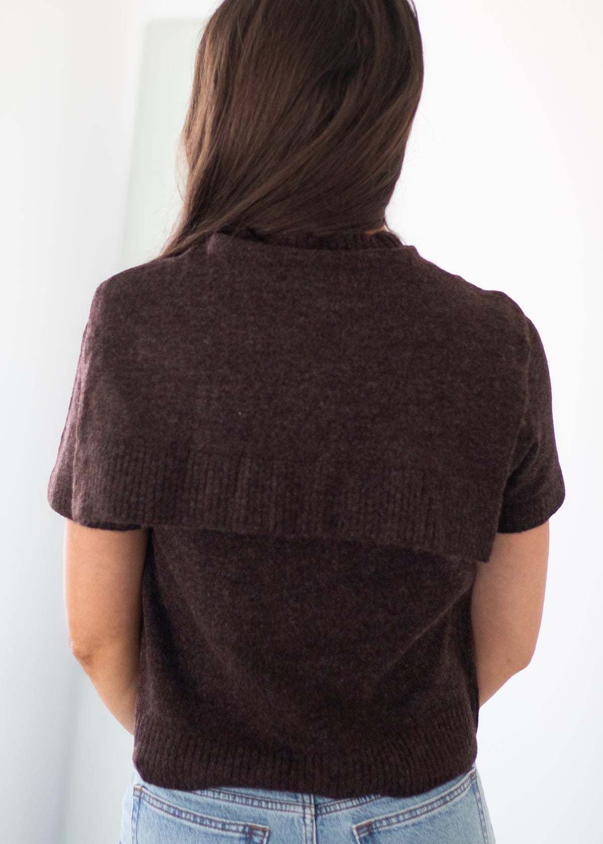 A woman viewed from behind wearing a dark brown short-sleeve knit top with a wide ribbed yoke and light blue jeans against a plain white background.