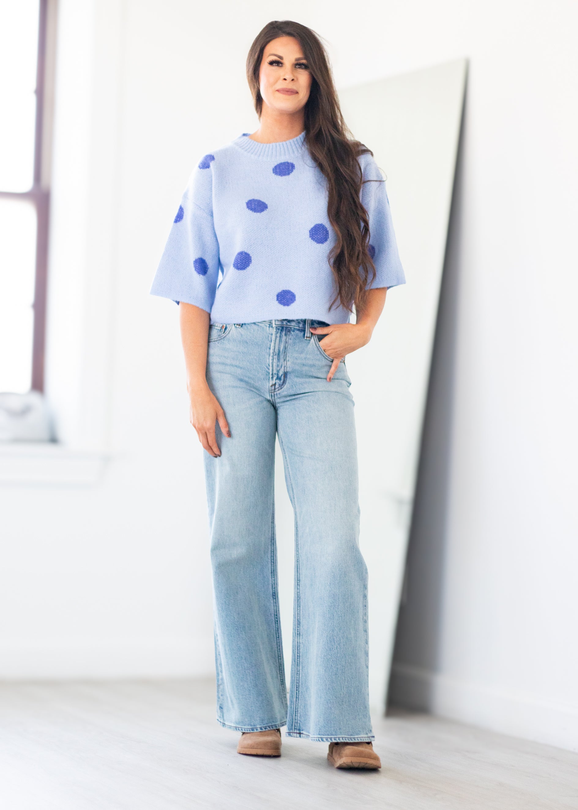 A woman modeling a light blue knit sweater with dark blue polka dots, paired with light-wash wide-leg jeans and brown shoes, standing in a bright white room.