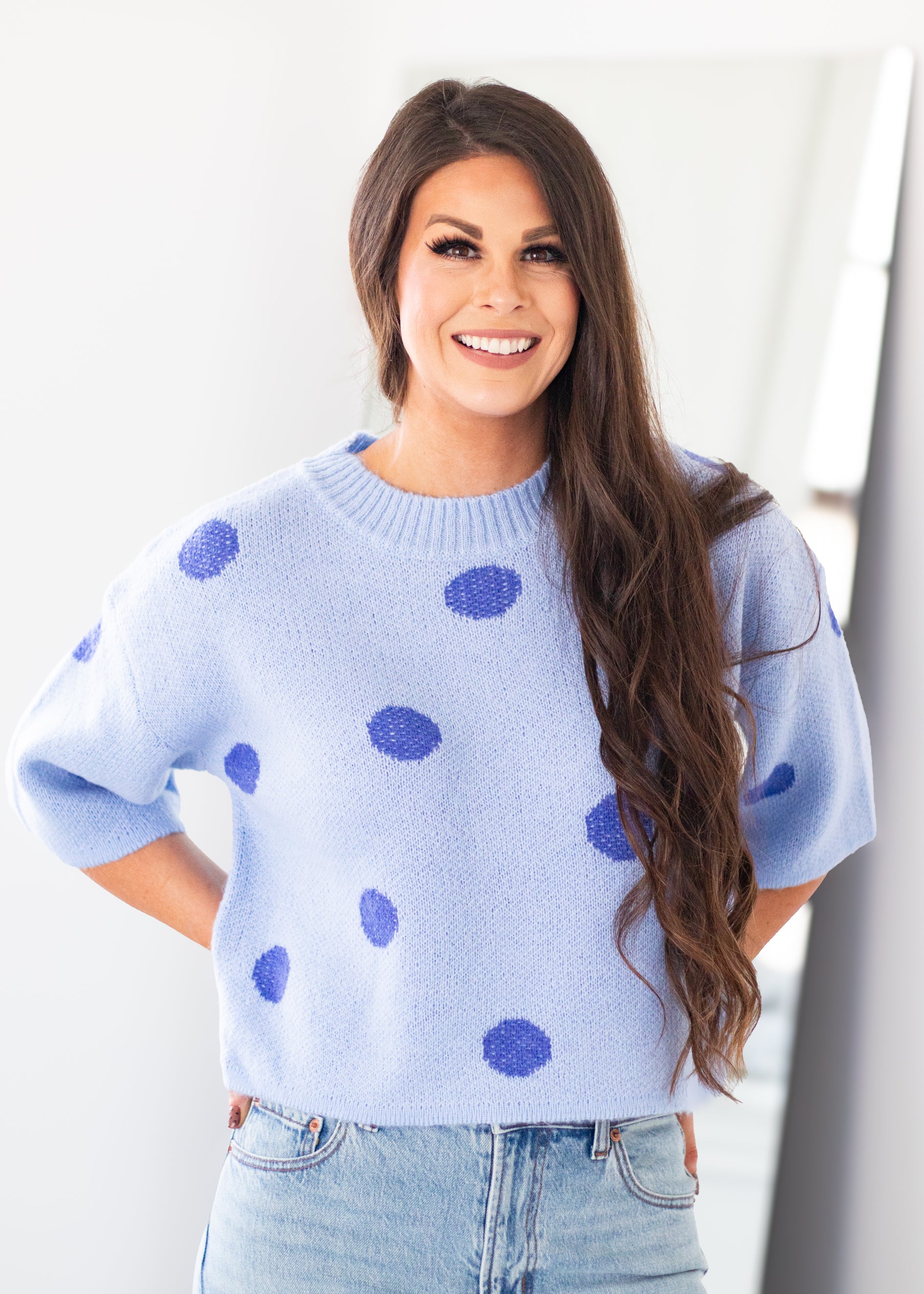A smiling woman with long brown hair wearing a light blue knit sweater with dark blue polka dots and light-wash blue jeans, standing against a bright white indoor background.