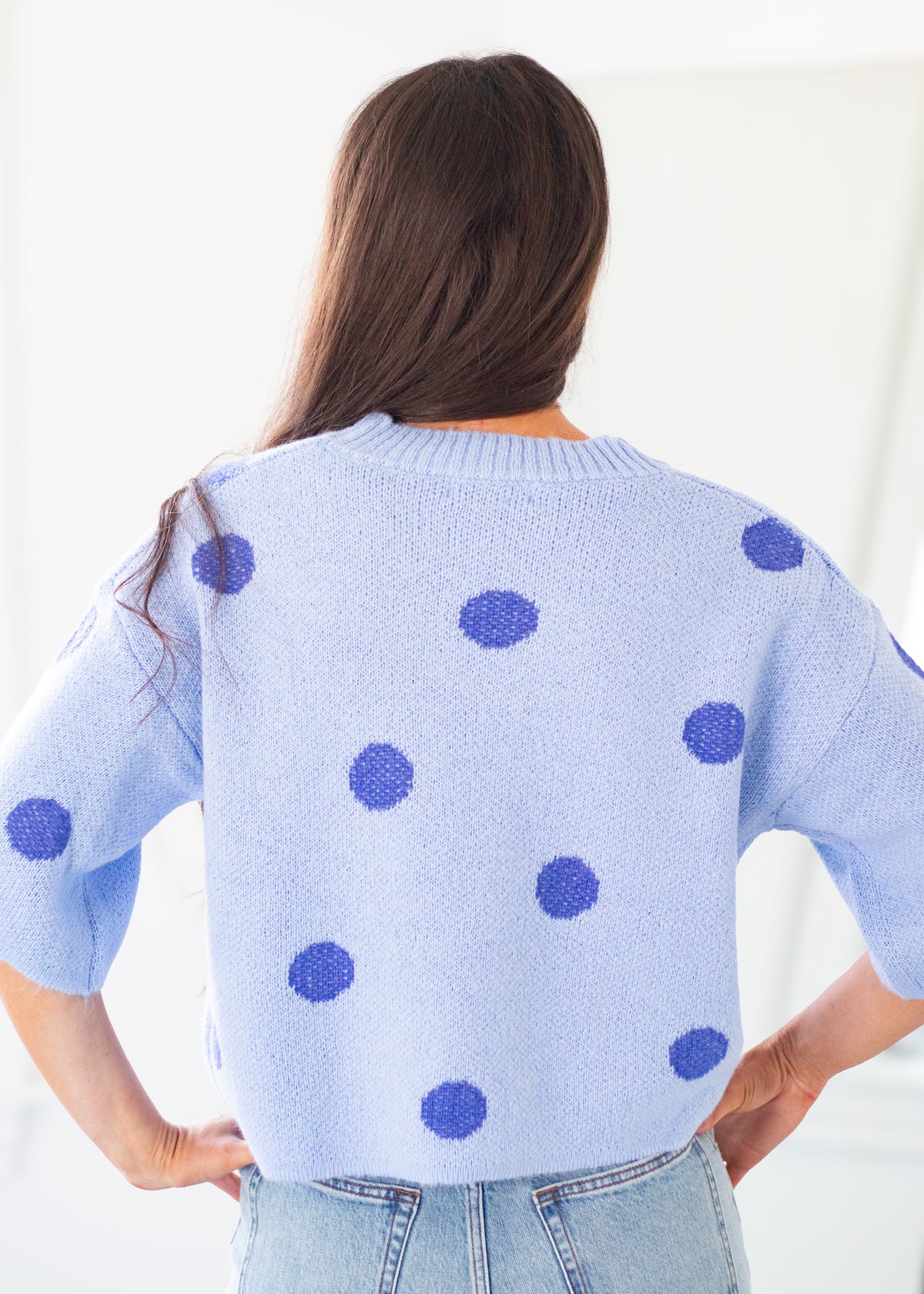 A woman viewed from behind wearing a light blue knit sweater with dark blue polka dots, paired with light-wash jeans in a bright indoor setting.