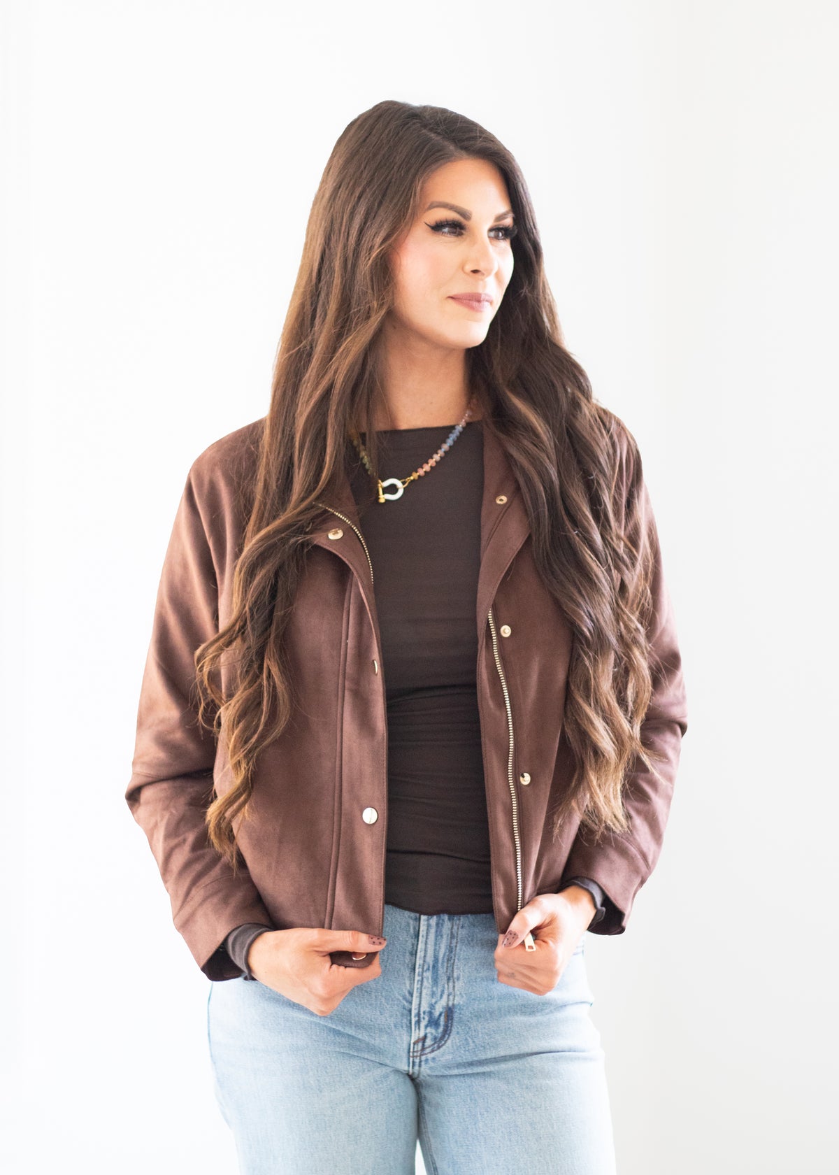 A woman modeling a brown faux-leather jacket over a dark brown top and light blue jeans, standing against a plain white studio background.