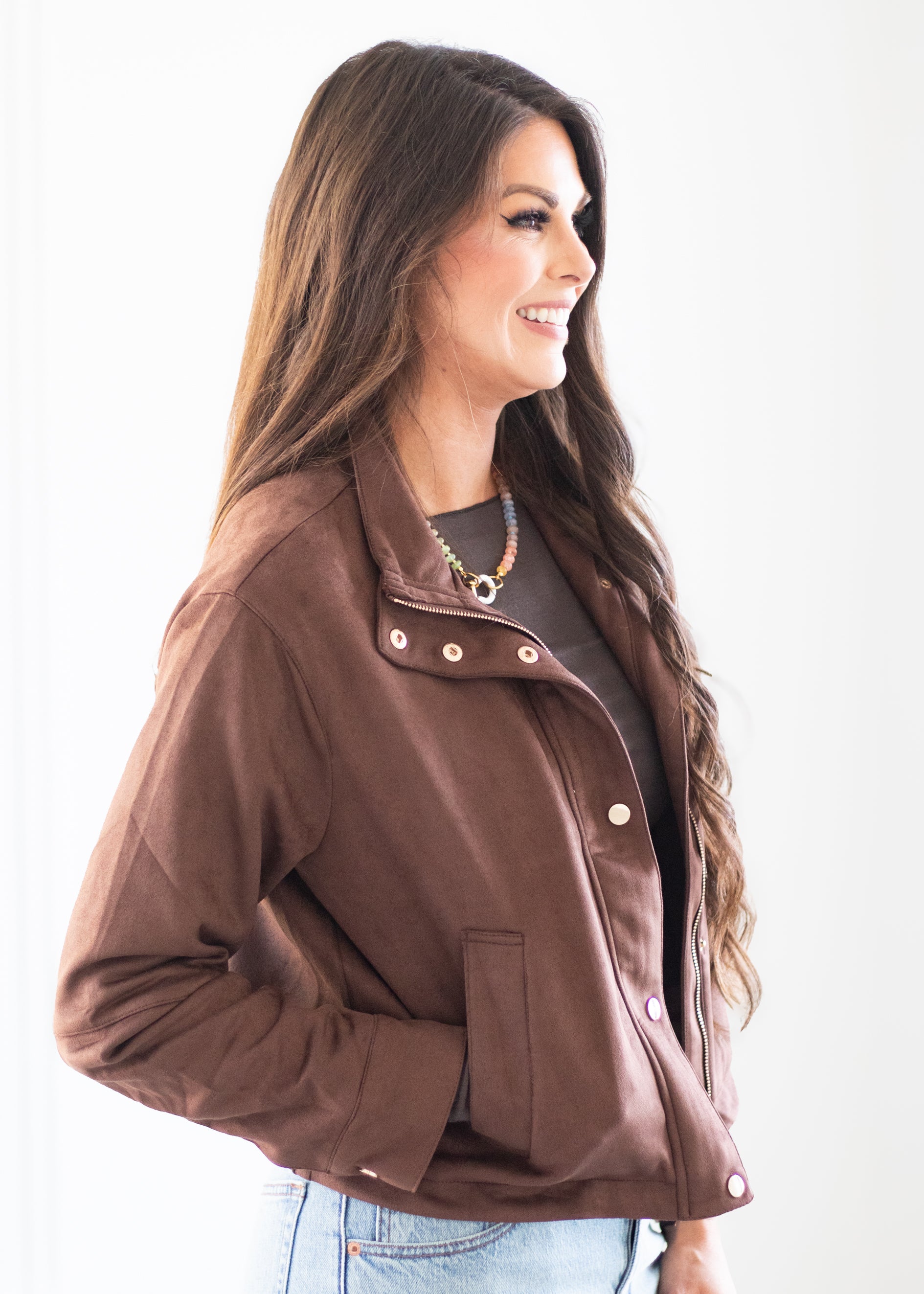 A woman wearing a brown cropped jacket with snap buttons and a pointed collar, styled over a dark top and light blue jeans, photographed against a plain white background.