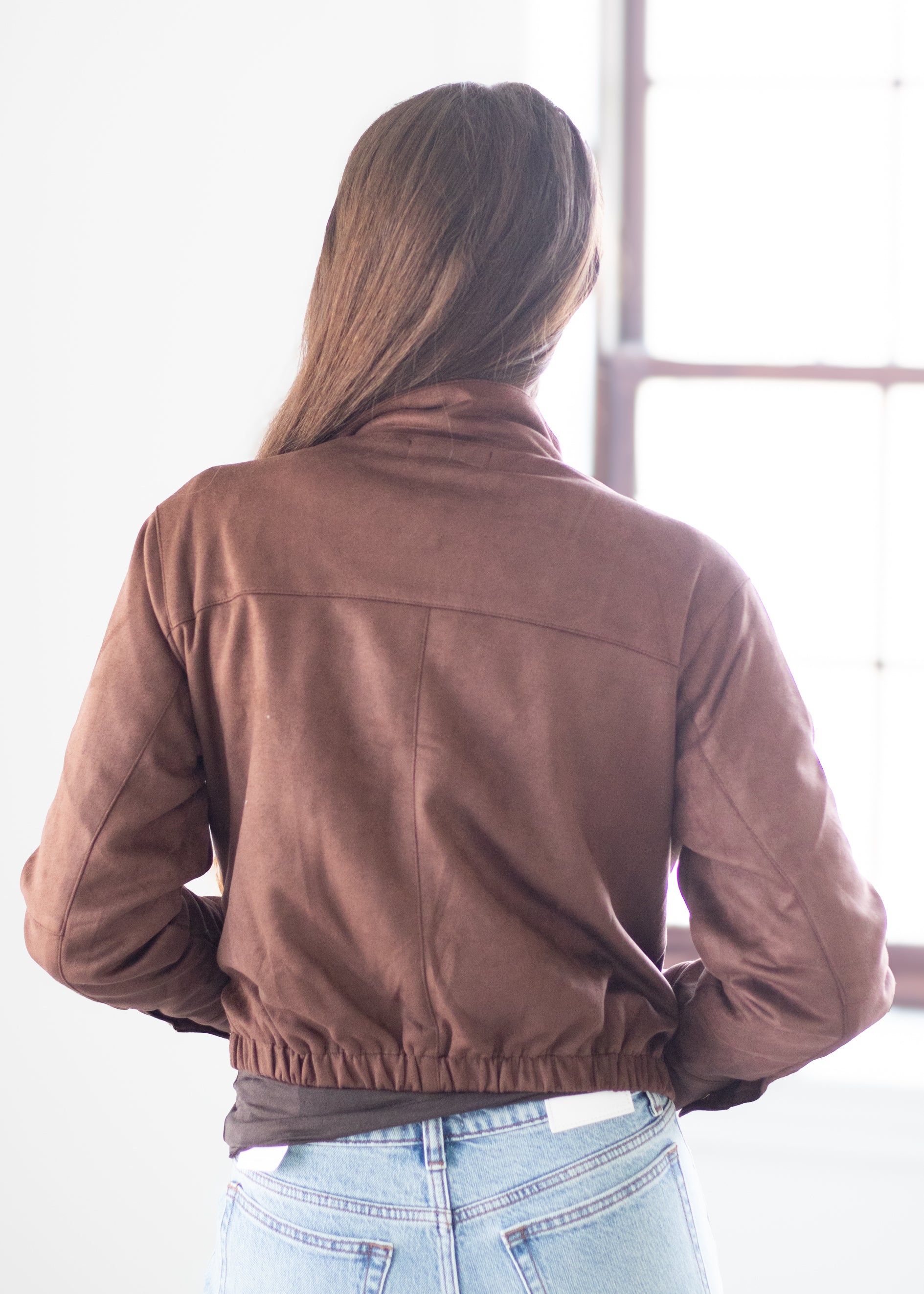 A woman viewed from behind wearing a brown suede-like cropped jacket and light blue jeans, standing in a bright indoor setting near a window.