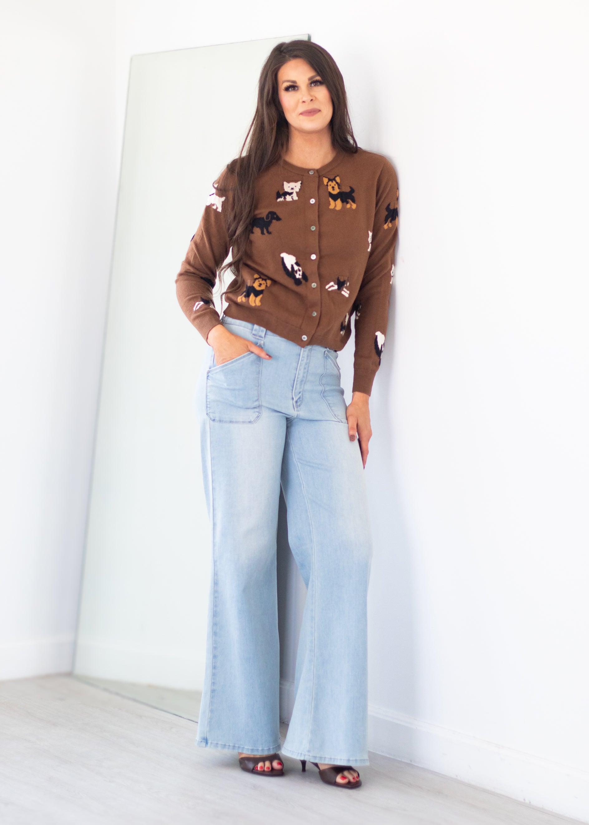 A woman stands in a bright room wearing a brown button-front cardigan with a multicolor animal print, light-wash wide-leg jeans, and black sandals.