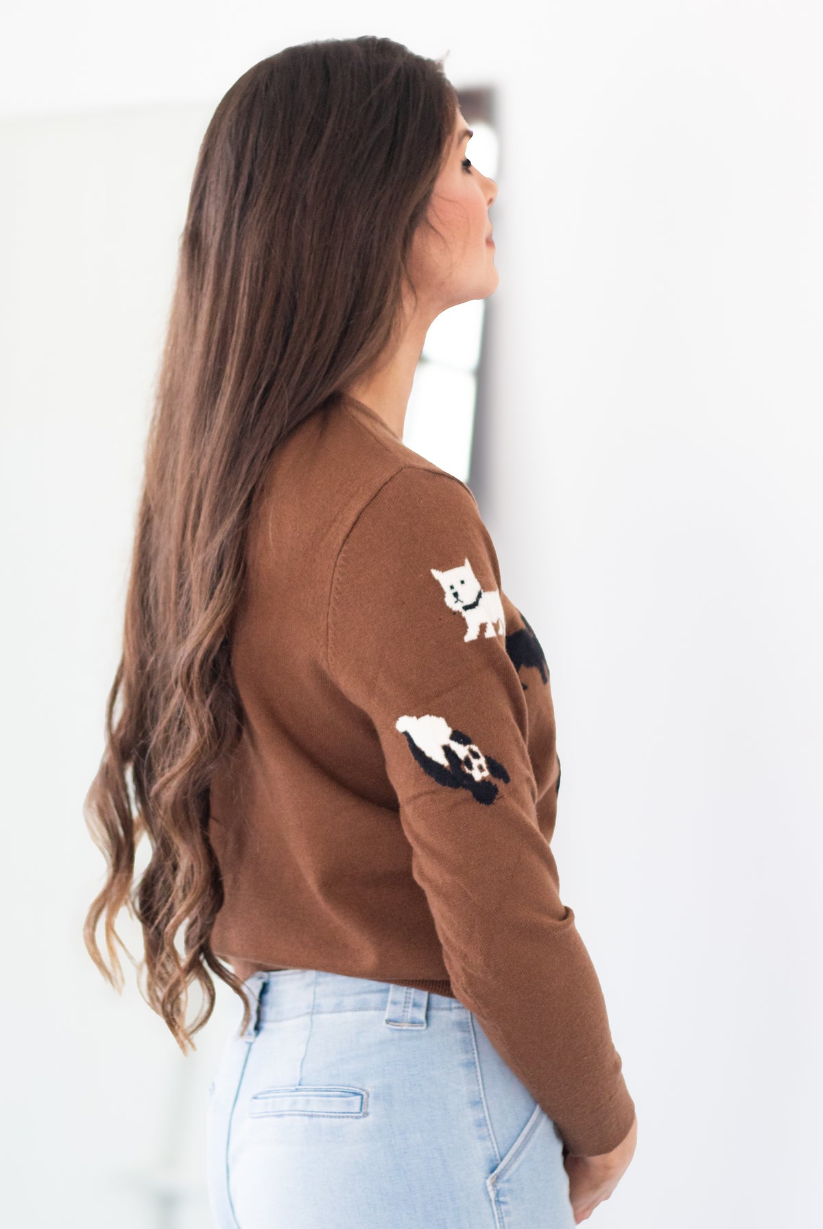 A woman with long brown hair is shown from a side-back angle wearing a brown knit sweater with embroidered cat and panda motifs on the sleeves, paired with light blue jeans against a bright white background.