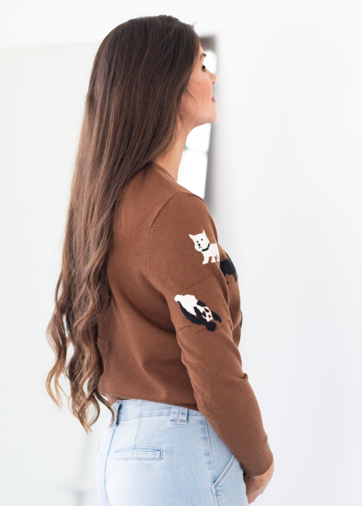A woman with long brown hair is shown from a side-back angle wearing a brown knit sweater with embroidered cat and panda motifs on the sleeves, paired with light blue jeans against a bright white background.
