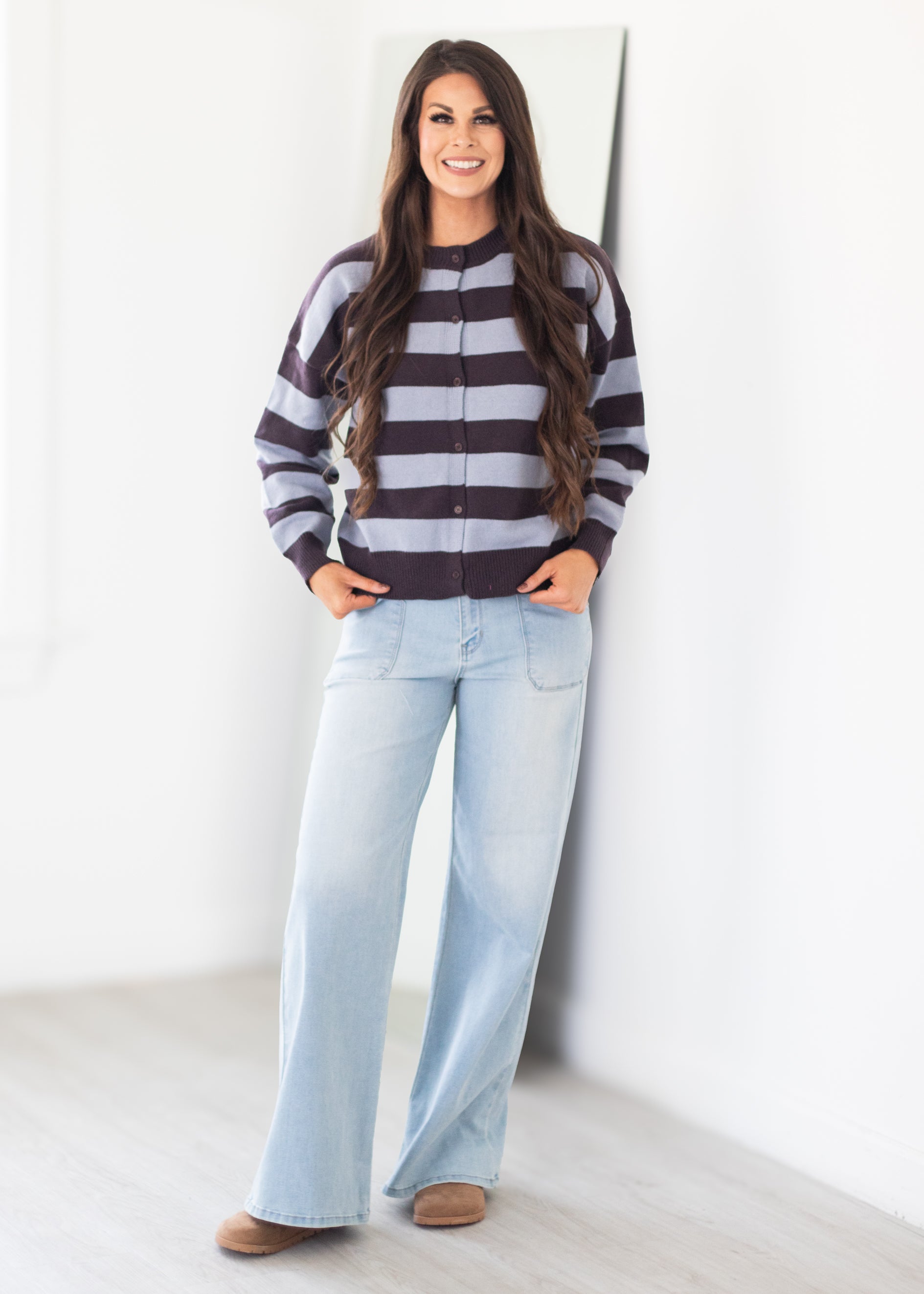 A woman modeling a dark brown and light gray striped zip-up sweater with wide-leg light blue jeans and brown shoes, standing against a white wall.