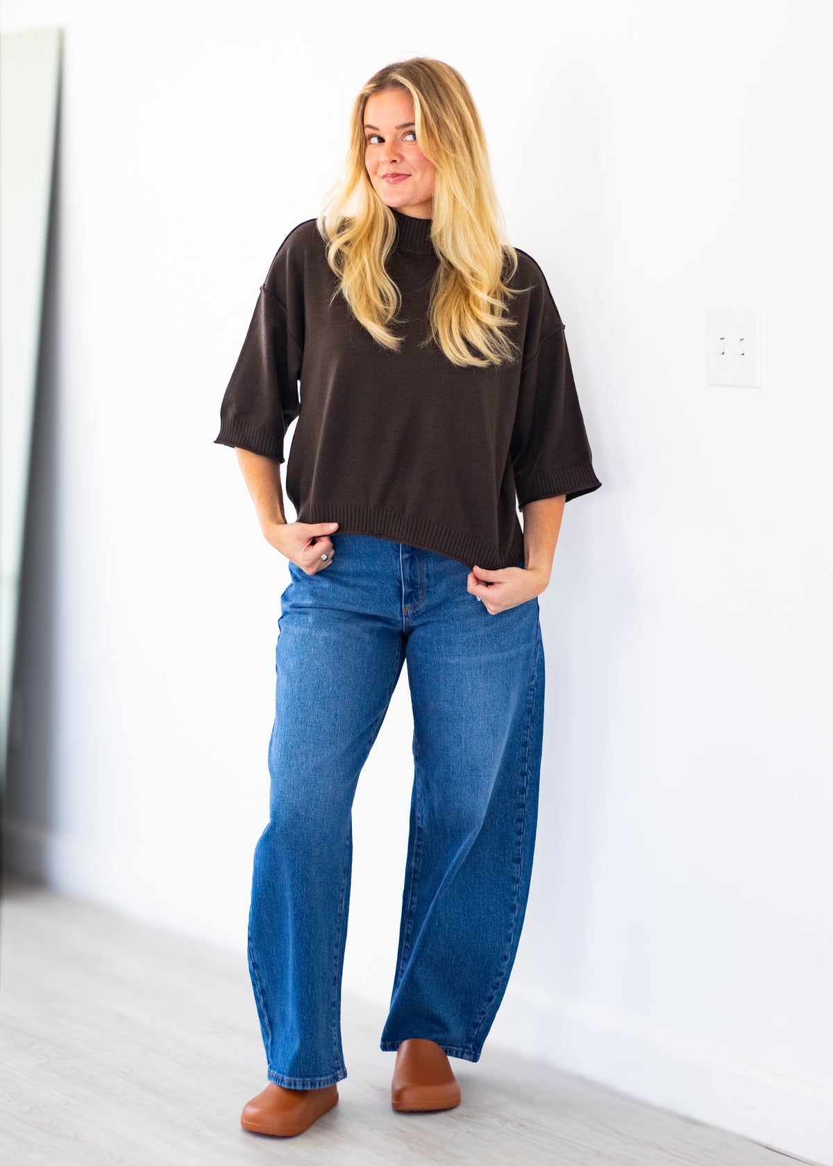 A woman stands indoors wearing a dark brown mock-neck tunic top, blue wide-leg jeans, and brown shoes against a white wall.