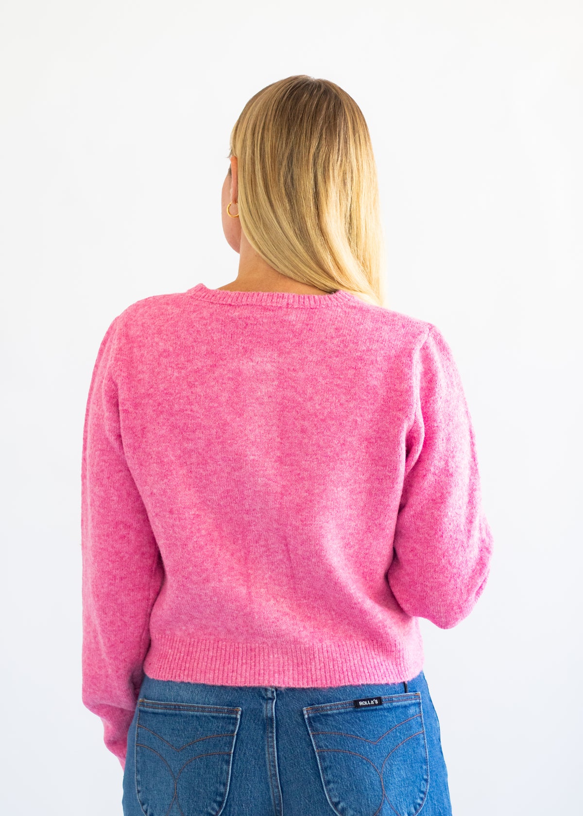 A woman viewed from behind wearing a bright pink knit sweater and blue jeans against a plain white background.