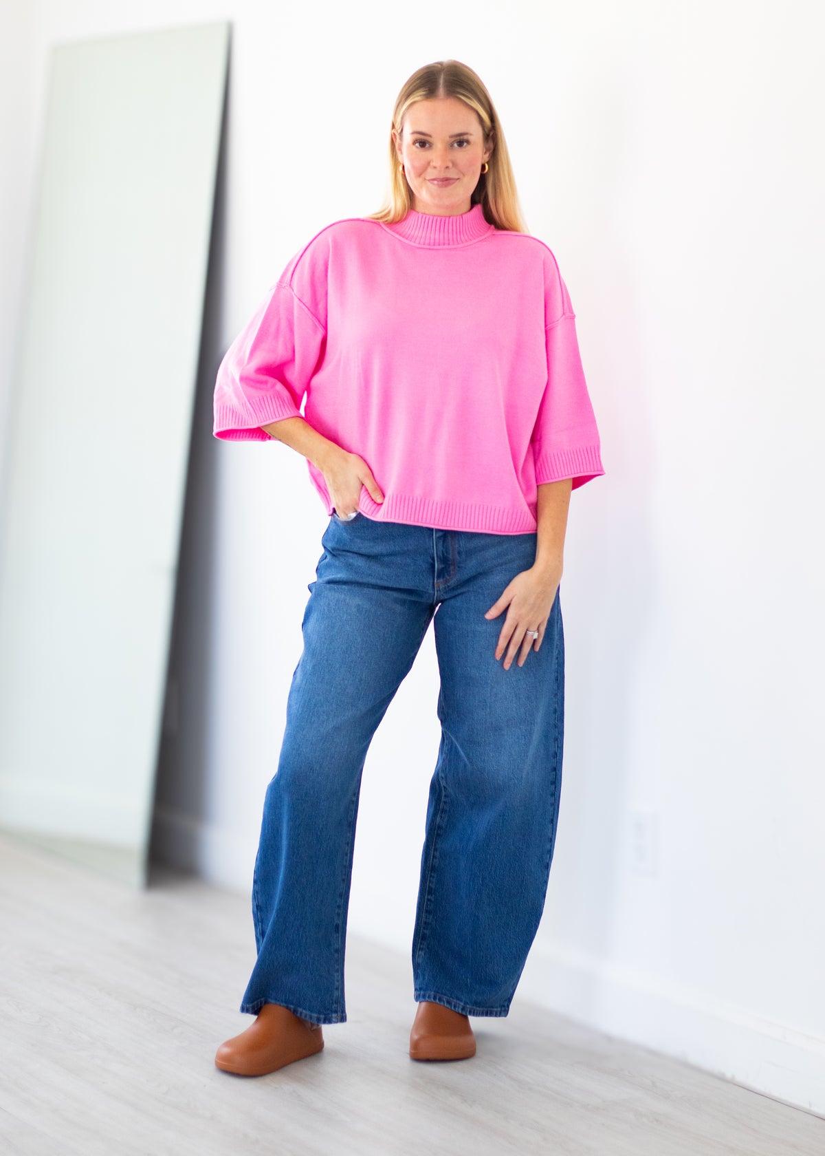 A woman modeling a bright pink mock-neck short-sleeve top with wide legs and blue jeans, standing in a minimalist room with a white wall and light wood floor.