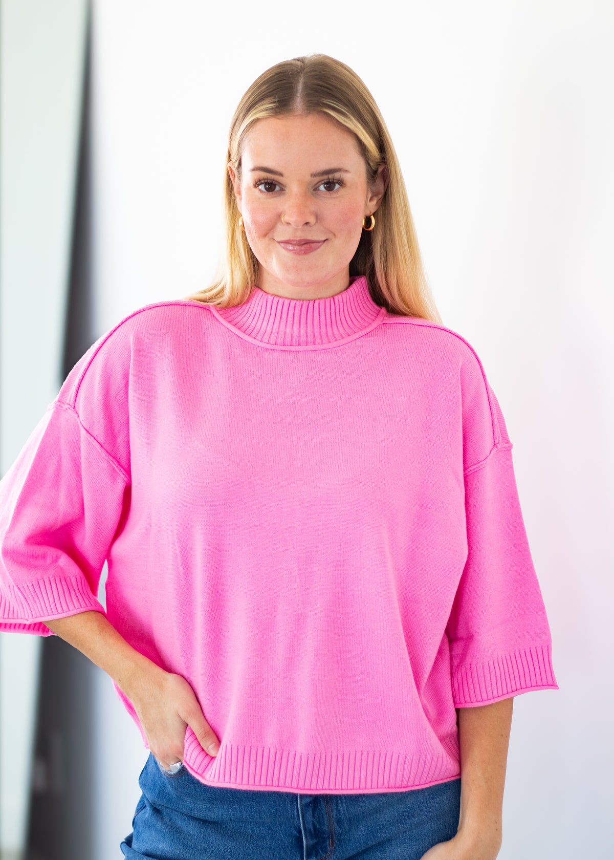 A woman wearing a bright pink mock-neck knit sweater with three-quarter sleeves and blue jeans, standing against a white wall.