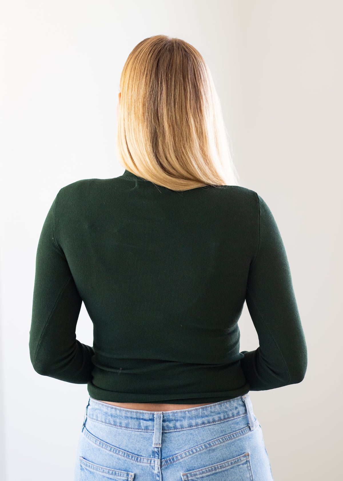 A woman viewed from behind wearing a dark forest green long-sleeve mock neck top and light blue jeans, standing against a plain off-white wall.