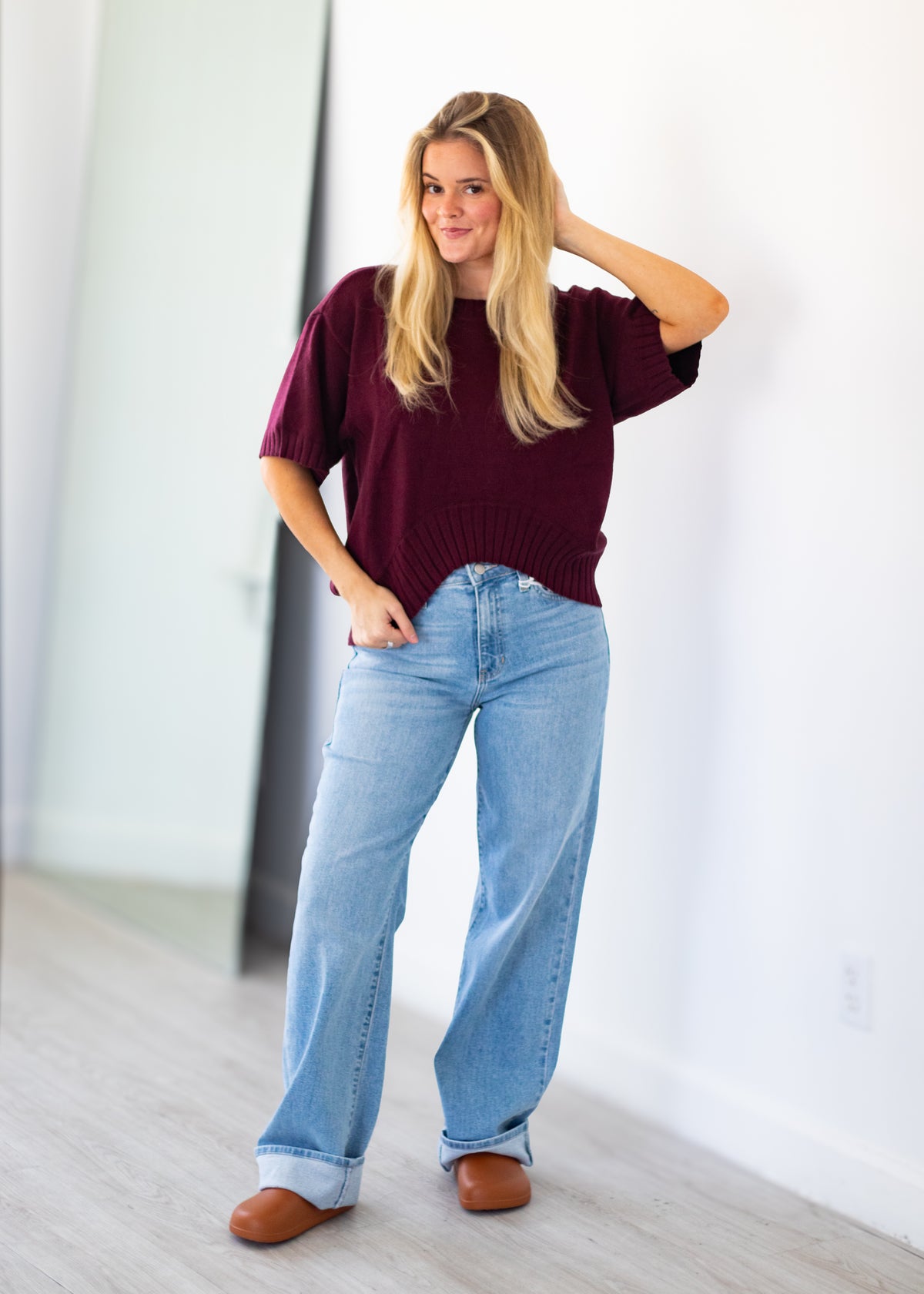 A woman modeling a short-sleeve burgundy knit sweater and light blue wide-leg jeans, standing in a bright indoor room with white walls and light wood flooring.