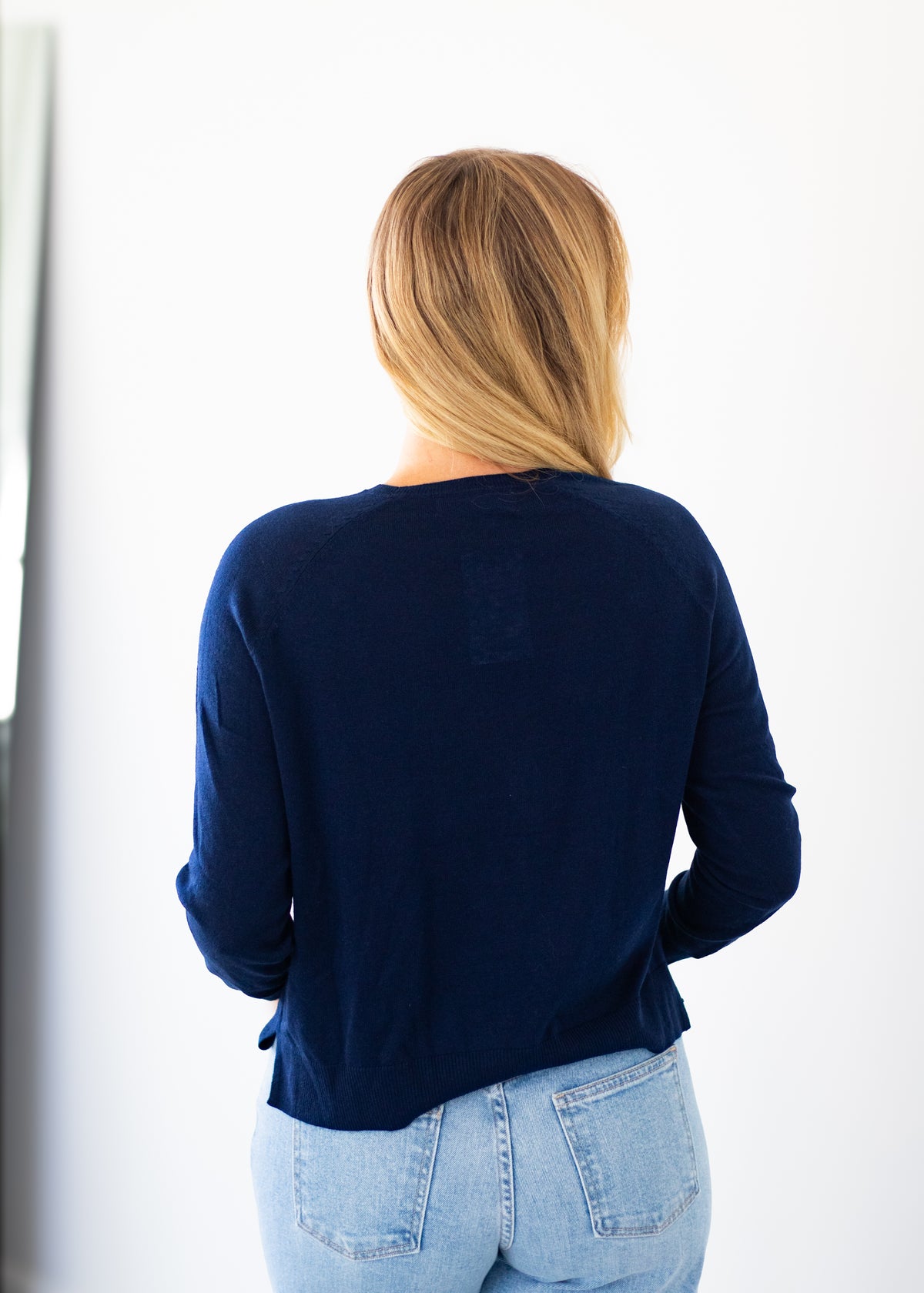 A woman viewed from behind wearing a dark navy blue long-sleeve top and light blue jeans against a bright white background.