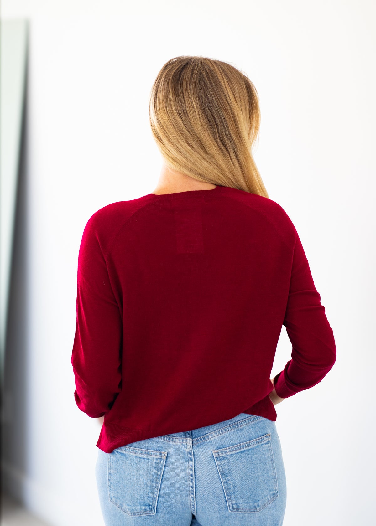A woman with blonde hair is shown from behind wearing a long-sleeve crewneck top in deep burgundy red and light wash blue jeans, standing against a plain white wall.