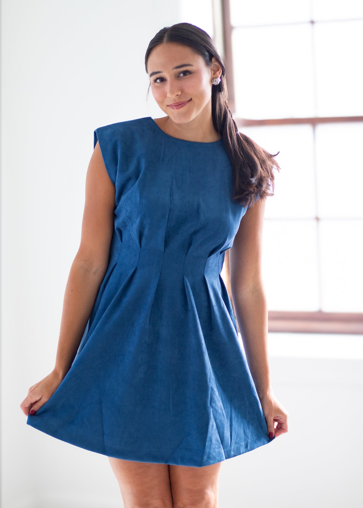 A woman wearing a sleeveless, blue A-line mini dress, standing in a bright room with a window in the background.