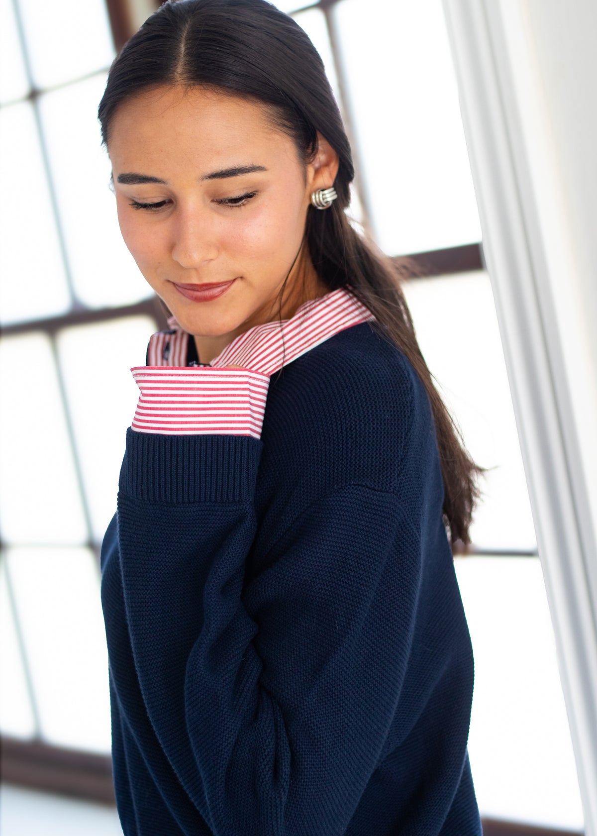 A woman wearing a navy blue ribbed knit sweater over a red and white striped shirt, standing in front of a bright window.