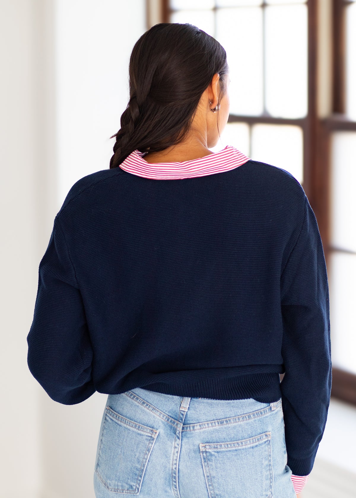 A woman viewed from behind wearing a navy blue knit sweater with a pink and white striped collar, paired with light blue jeans in a bright indoor setting.