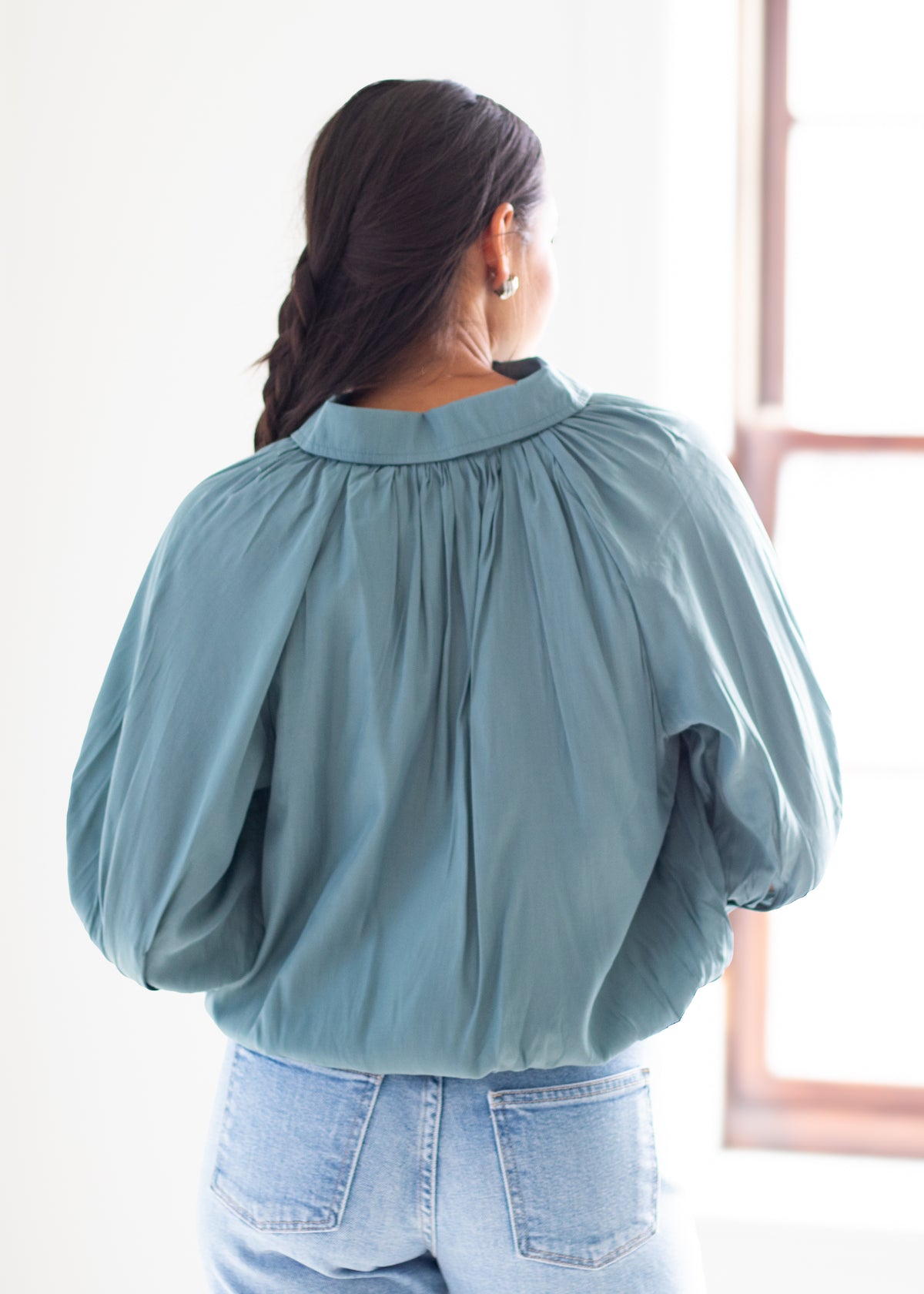 A woman viewed from behind wearing a dusty blue long-sleeve blouse with a gathered, pleated back and light-wash blue jeans, standing in a brightly lit room.
