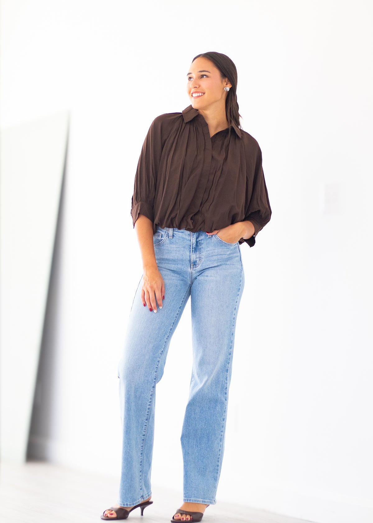 A woman modeling a dark brown pleated button-down shirt and light-wash blue jeans, standing against a bright white wall.