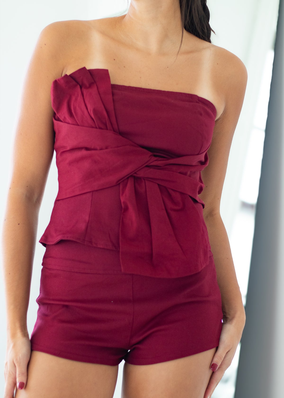 A woman wearing a strapless wine-red romper with a large twisted bow detail on the chest and matching fitted shorts.