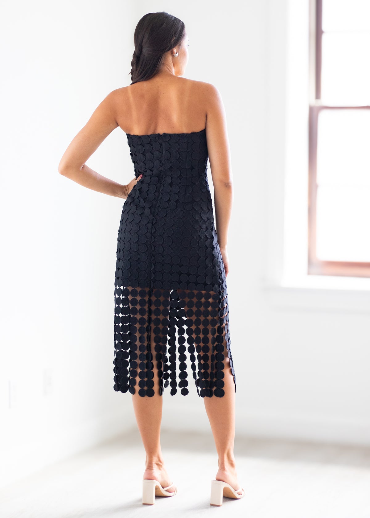 A woman shown from the back wearing a strapless black crochet midi dress with a textured pattern and a sheer lower section with hanging crochet details, paired with white block-heel sandals in a bright white room.