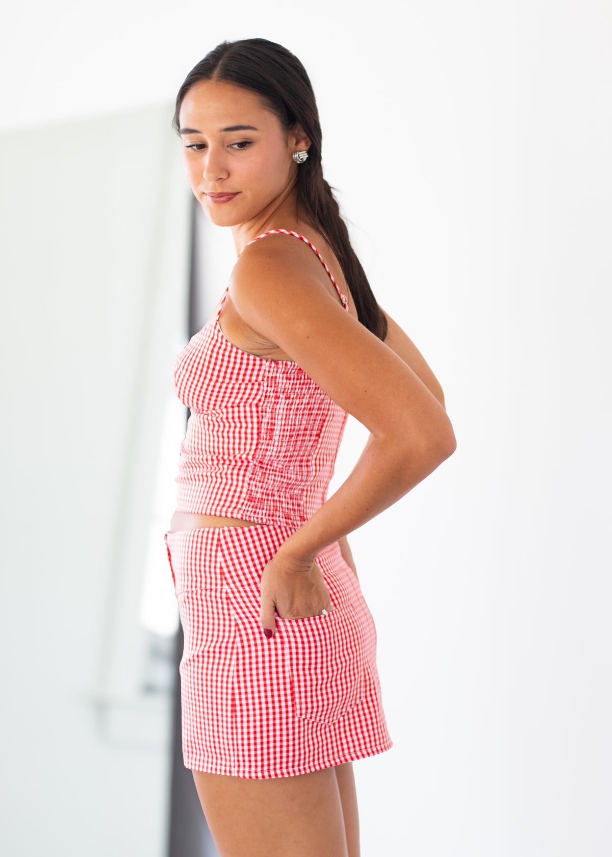 A woman modeling a red and white gingham two-piece set consisting of a sleeveless crop top and matching shorts, shown from a side-back angle against a bright white studio background.