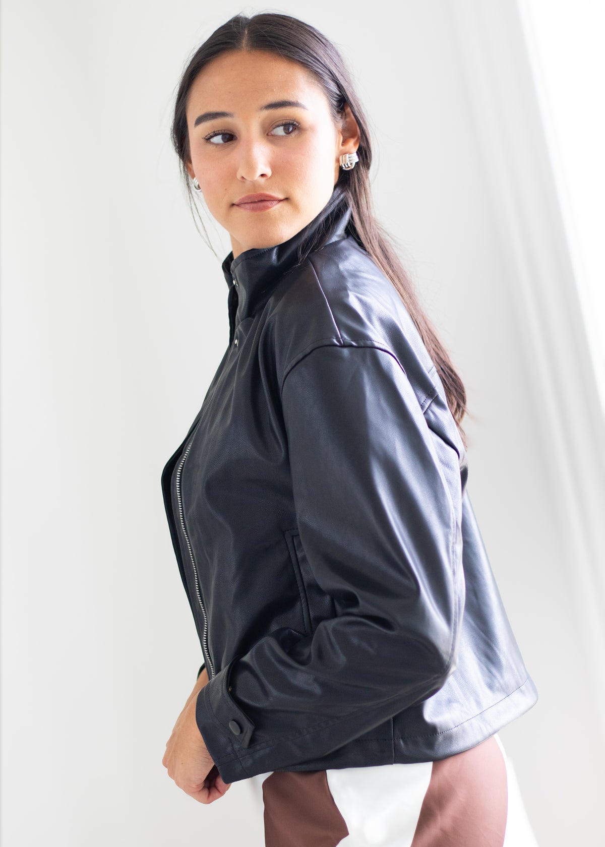A woman wearing a black faux-leather jacket, photographed from a side-back angle against a bright, neutral indoor background.