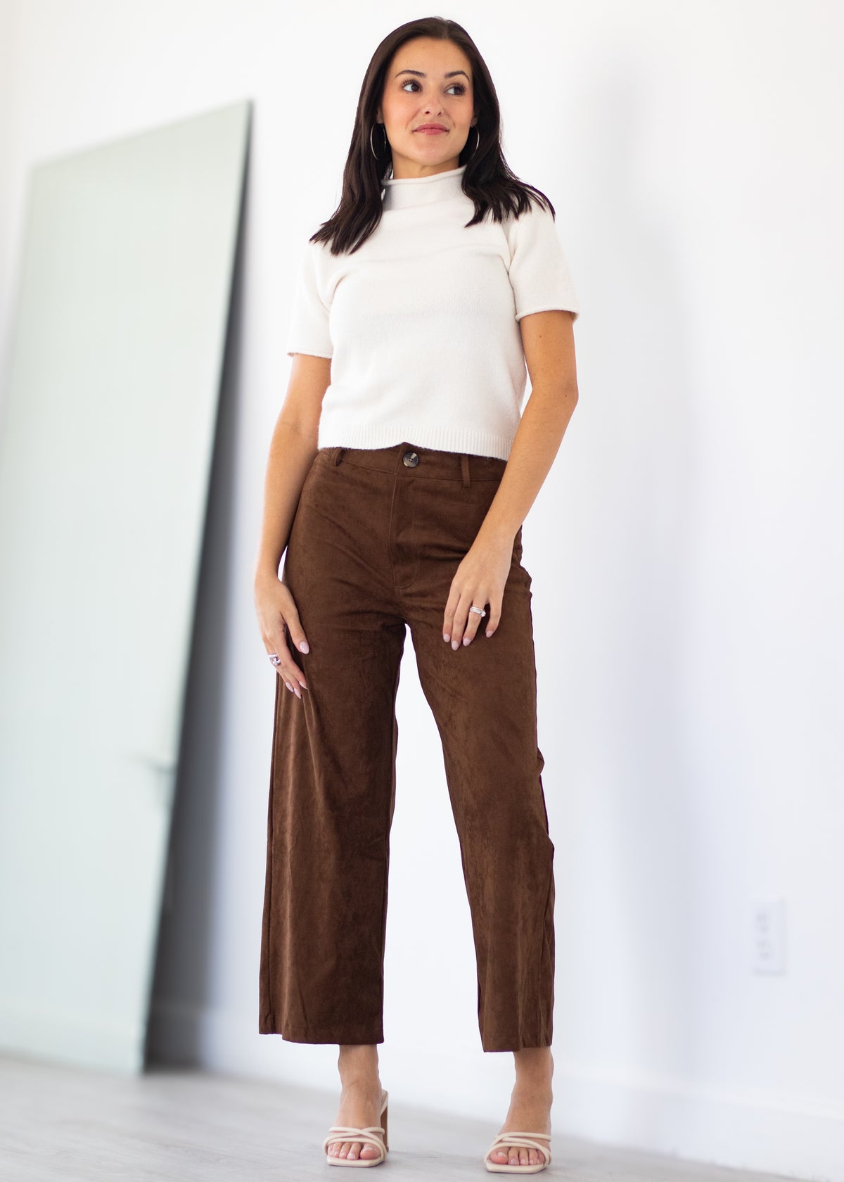 A woman modeling a white short-sleeve mock neck top and brown wide-leg cropped pants, standing in a bright, minimalist room with a large floor mirror.