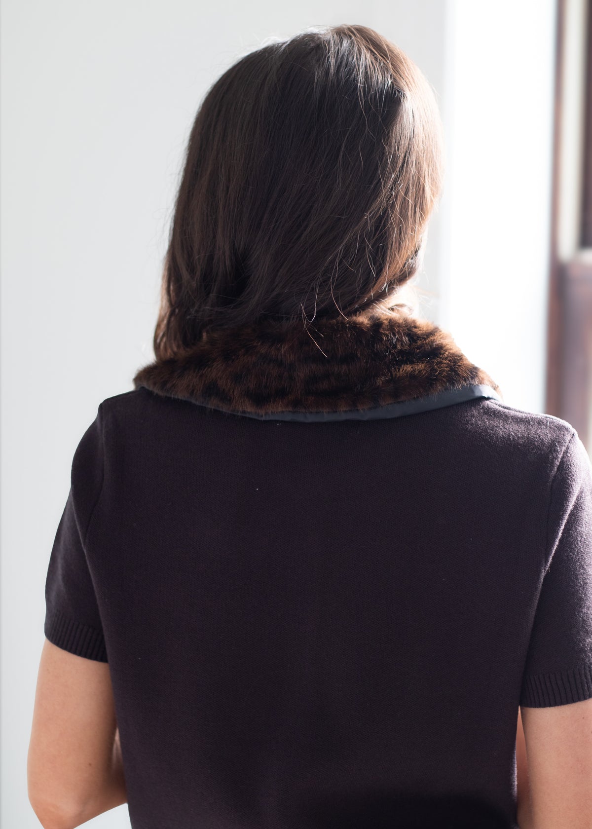 A woman viewed from behind wearing a dark brown faux-fur collar trim on a short-sleeve black knit top.